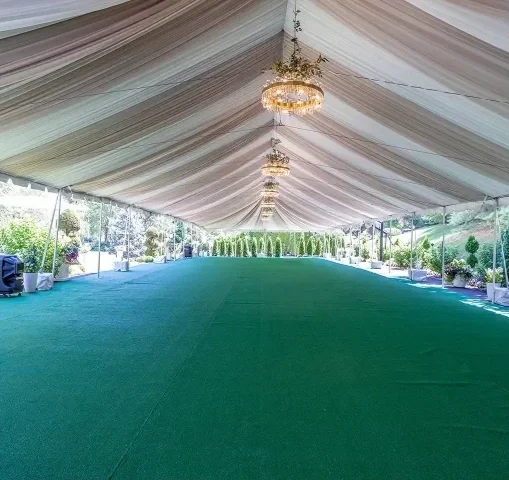 Large outdoor event tent with draped ceiling, hanging chandeliers, and a green carpeted floor, surrounded by plants and trees.