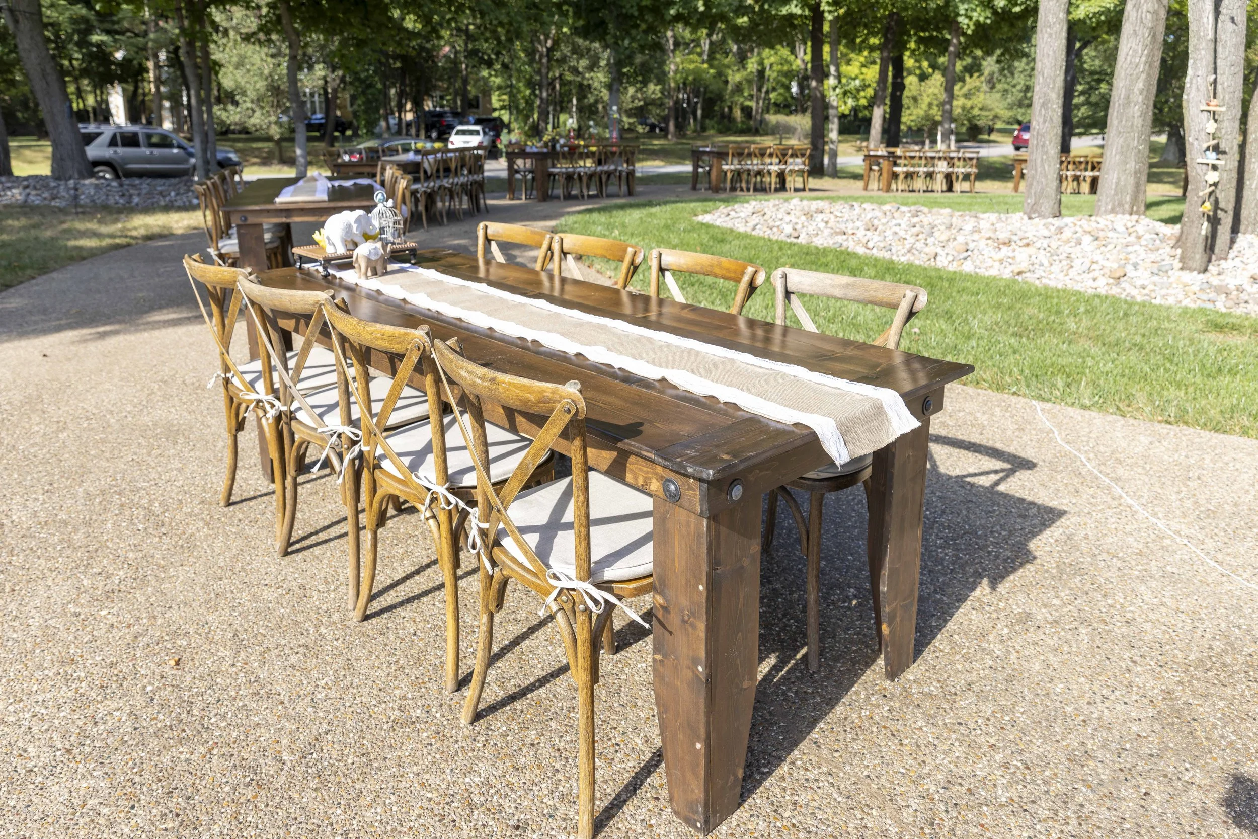 Outdoor gathering space with long wooden table and chairs, decorated with a table runner and small animal figurines, set in a park with trees and cars in the background.
