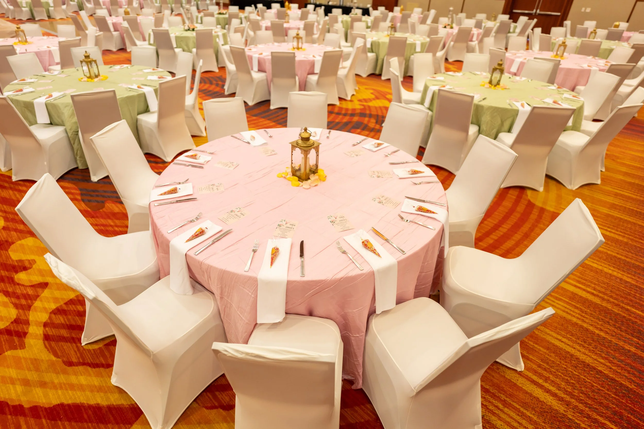 Banquet hall with round tables covered in pink and green tablecloths, each set with utensils, napkins, menus, and small lantern centerpieces. White chairs surround the tables on a patterned carpet.