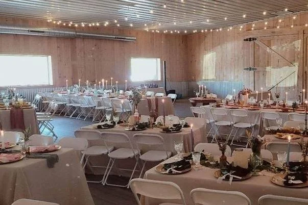 Rustic barn wedding reception decorated with round tables, white folding chairs, floral centerpieces, candles, and string lights