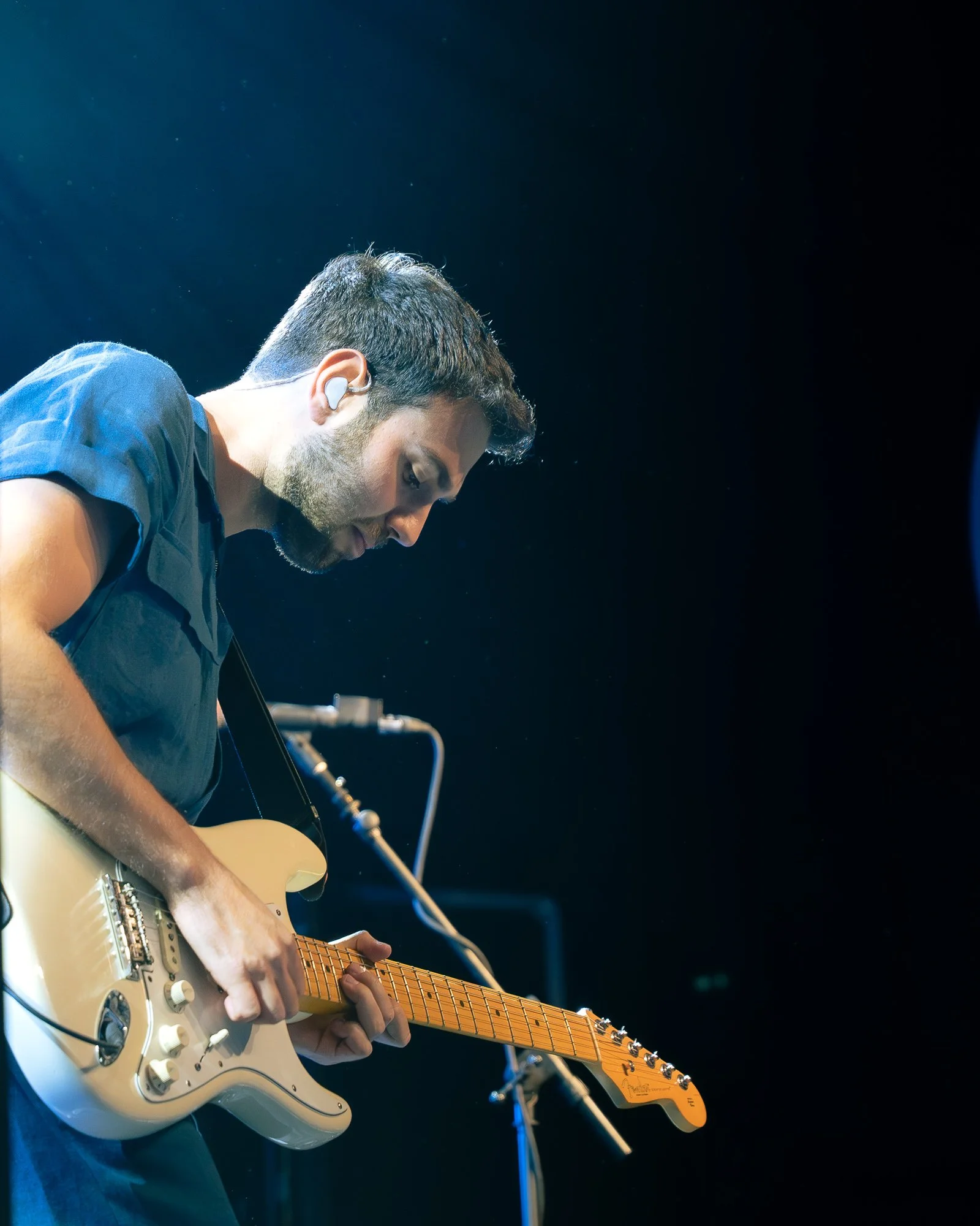 A male guitarist playing an electric guitar on stage, wearing a dark shirt, with a microphone stand nearby, under stage lighting.