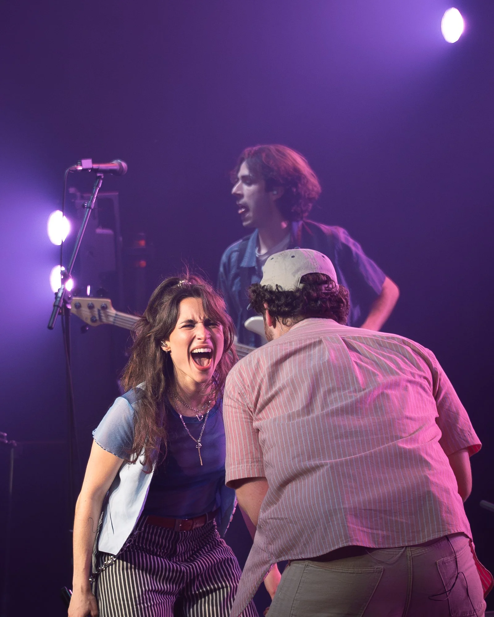 A lively concert scene with a woman singing passionately into a microphone, a man facing her, and a musician in the background playing a bass guitar on stage with purple and pink stage lighting.