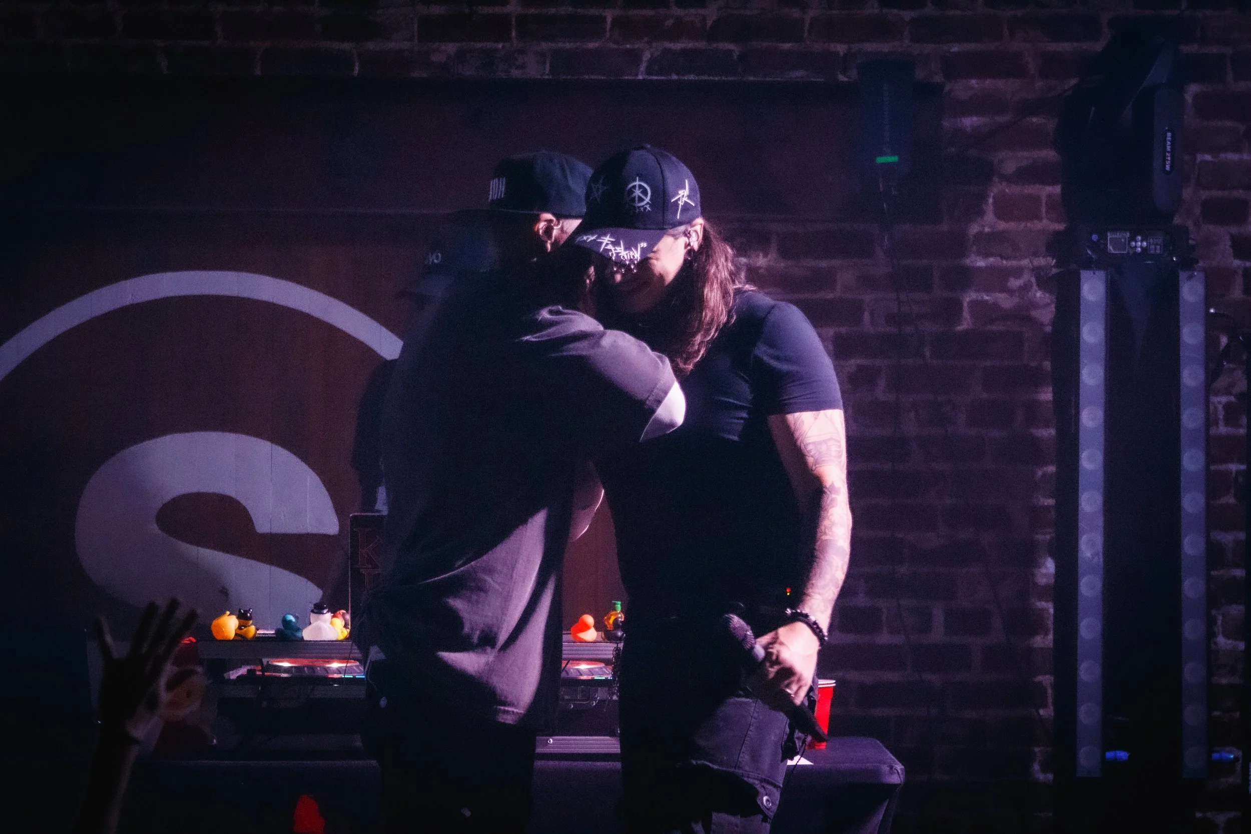 Two people on stage with a brick wall background, one holding a microphone, during a performance or karaoke night.