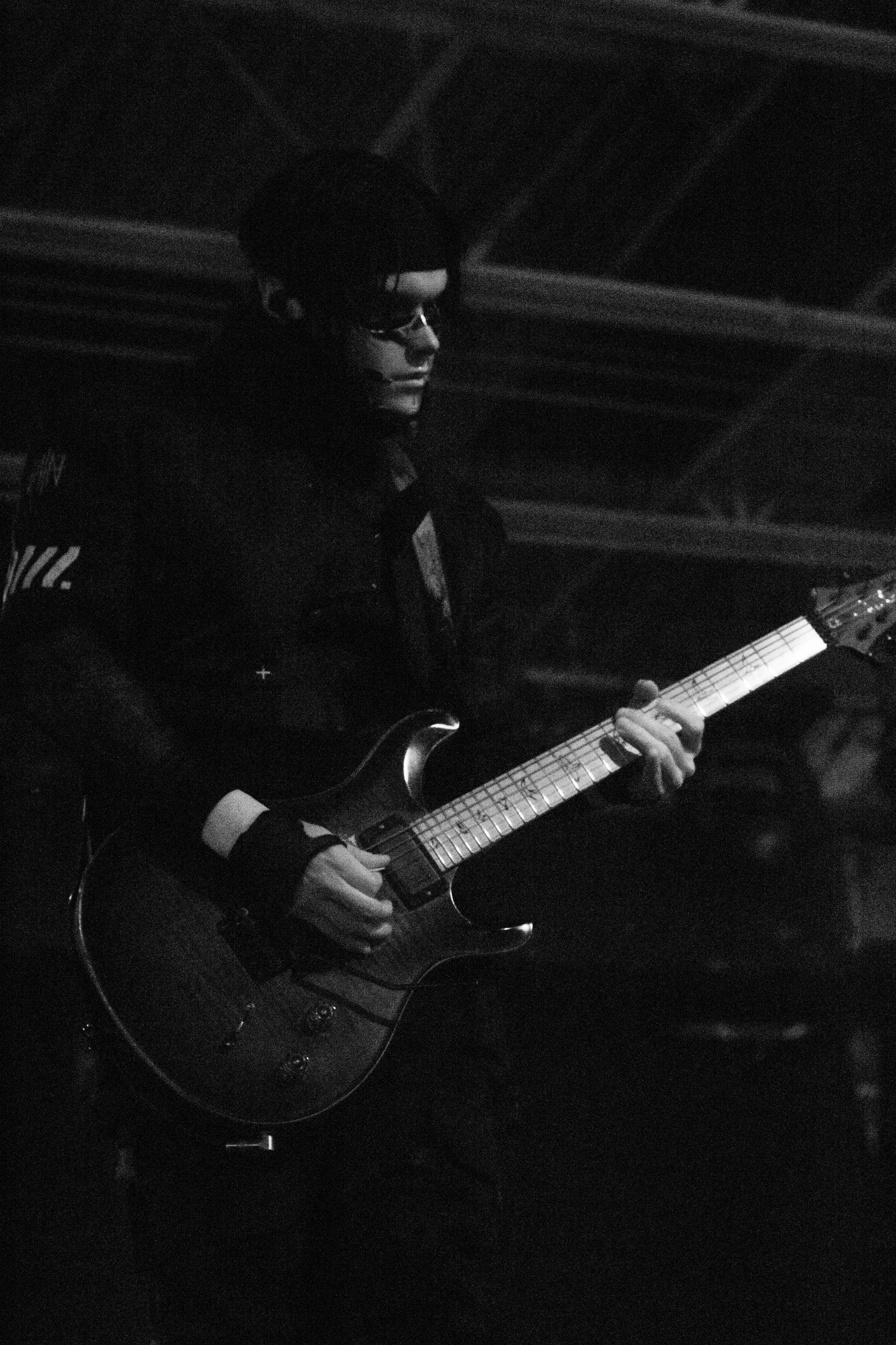 A person playing an electric guitar in a dark setting, wearing sunglasses and dark clothing.