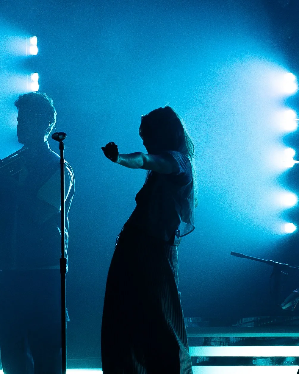 Silhouettes of two performers on stage with blue lighting, one in front of a microphone, the other extending an arm.