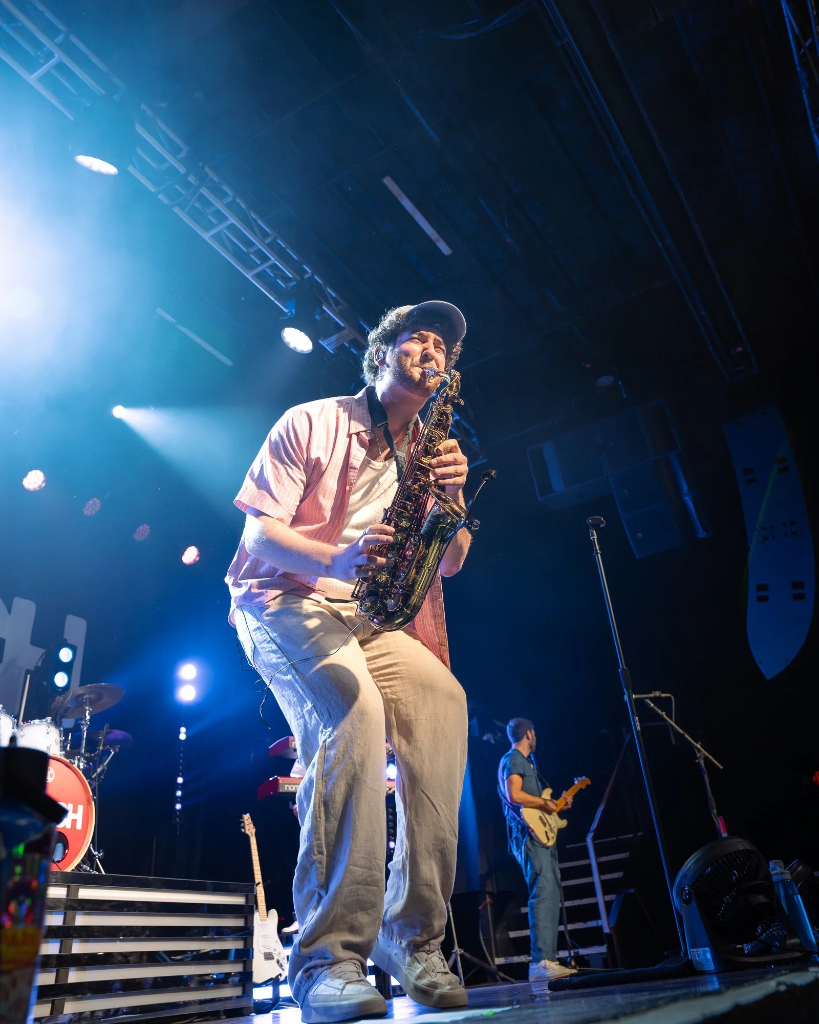 A man playing a saxophone on stage during a concert, with a guitarist in the background and stage lights illuminating the scene.