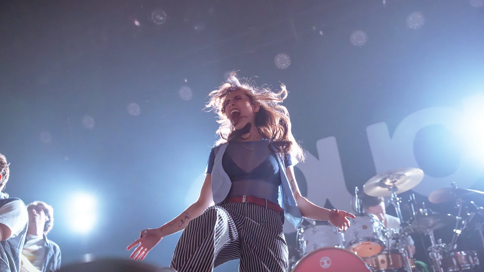 A female singer performing passionately on stage with her hair flying, wearing a sheer top, striped pants, and a blue jacket, with band members and drums in the background, illuminated by stage lights.