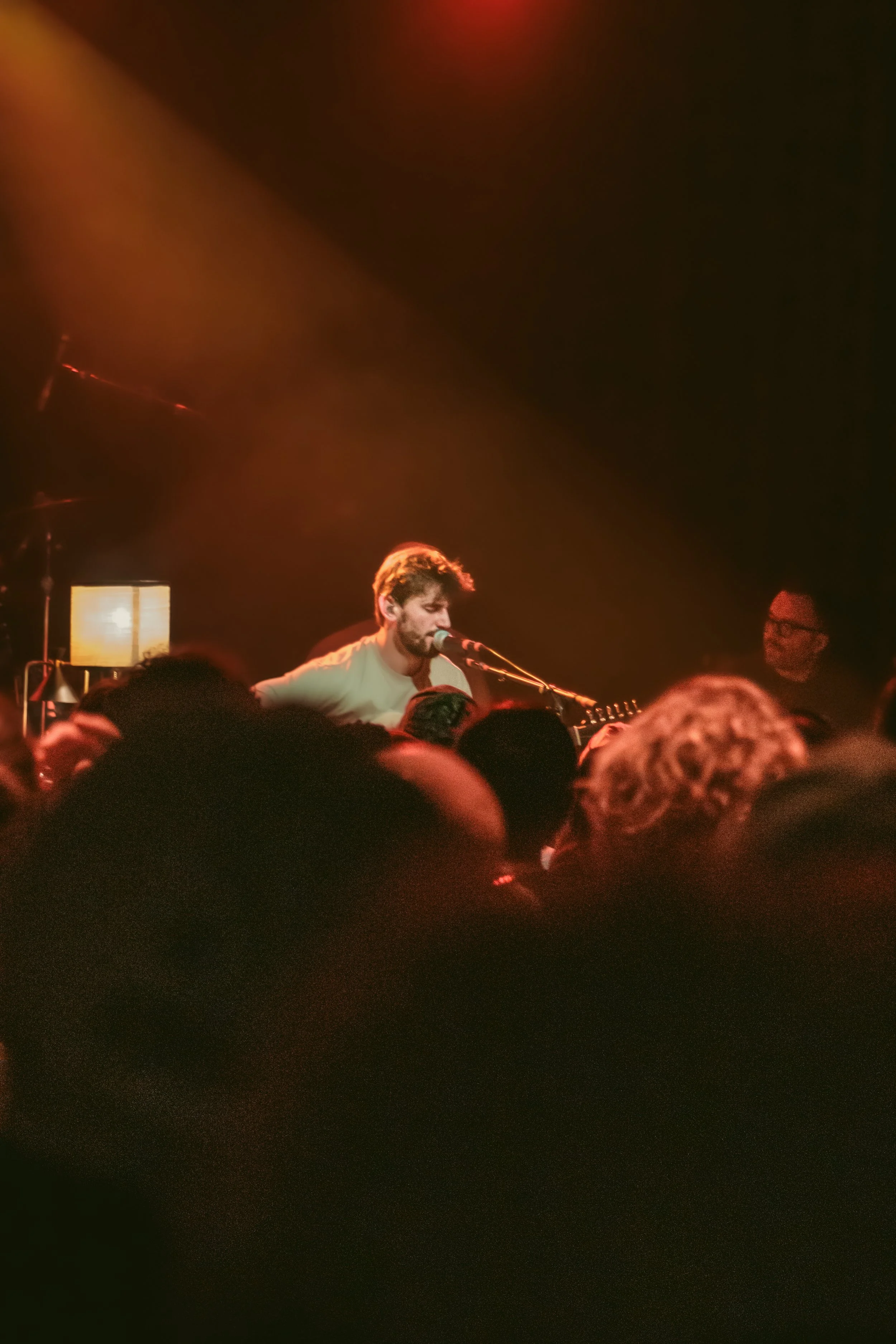 A musician playing a guitar and singing into a microphone on stage, with an audience in the foreground and warm stage lighting.