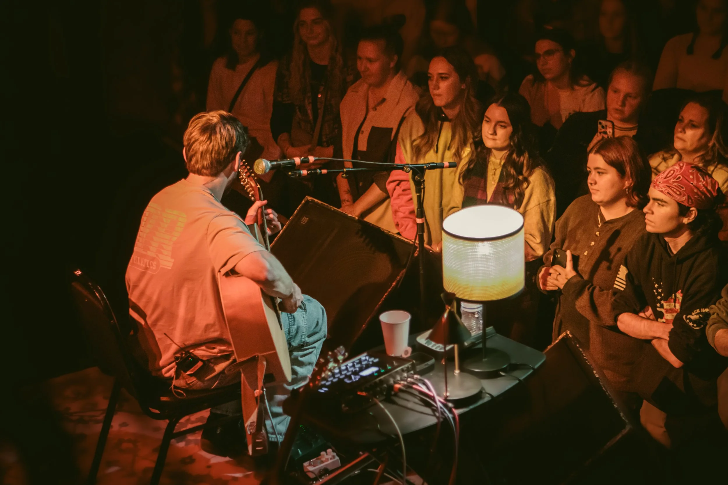 Person playing guitar and singing in front of a seated audience on a dimly lit stage with a lamp and audio equipment on a table.