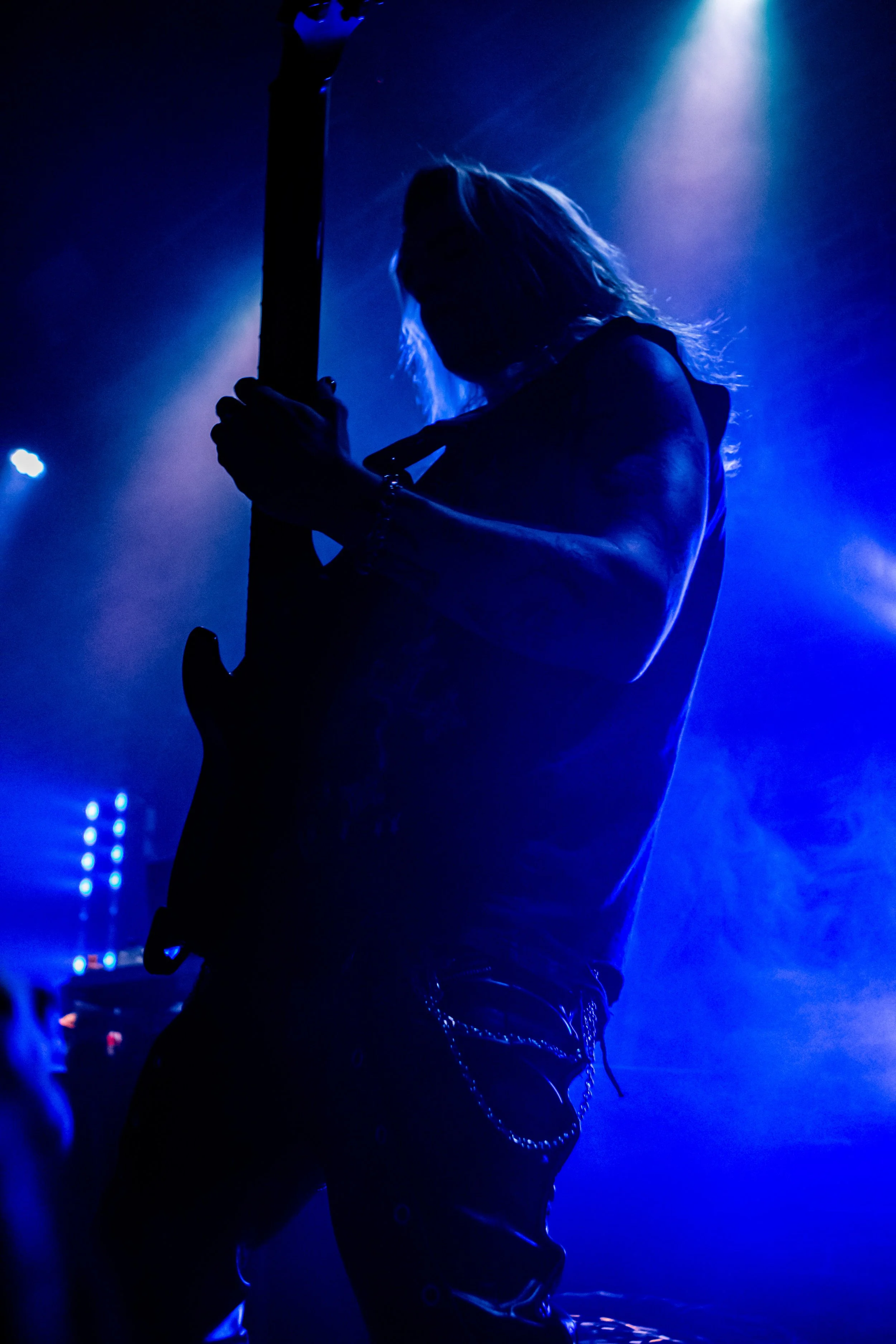 A person playing an electric guitar on stage during a concert, illuminated by blue lighting with a smoky background.