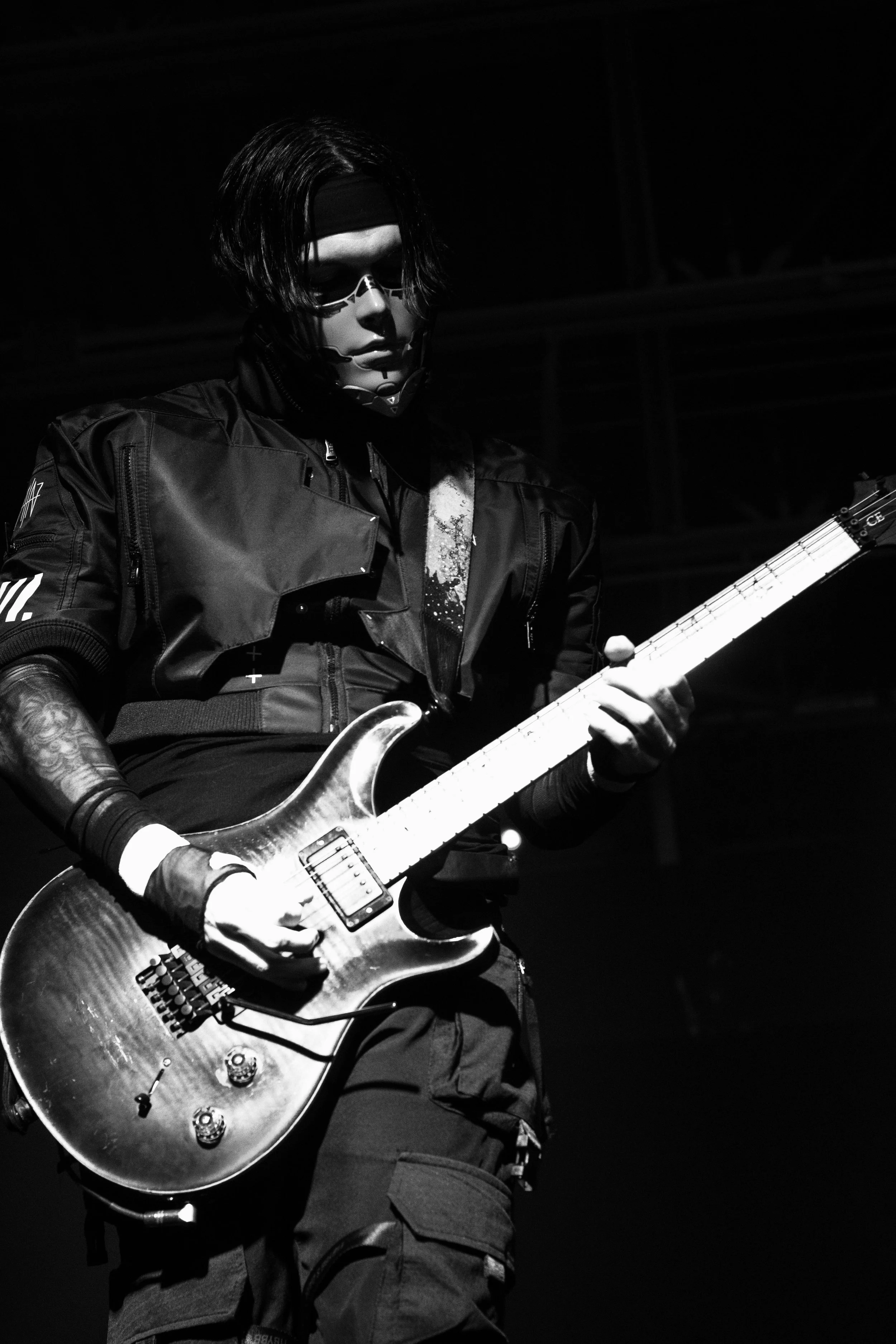 A musician playing an electric guitar on stage, dressed in dark clothing and wearing sunglasses and a headband, in black and white.