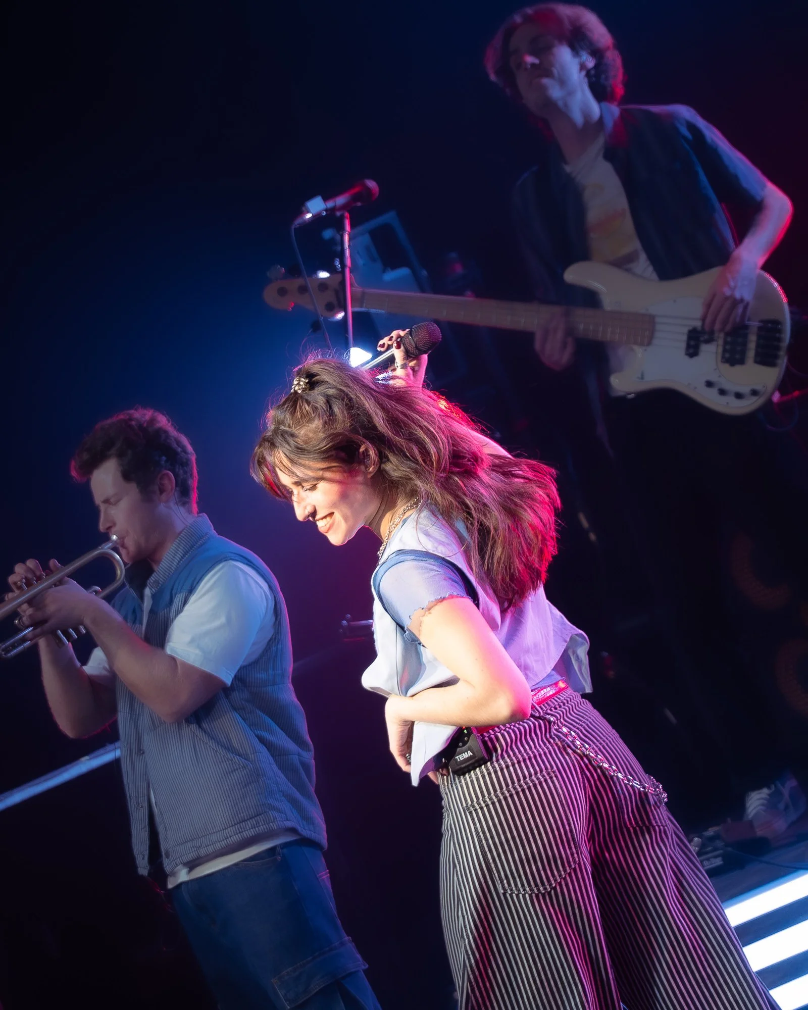 Young woman with wavy hair smiling and dancing on stage, holding a microphone, with a man playing trumpet beside her and a person in the background playing bass guitar under colorful stage lights.