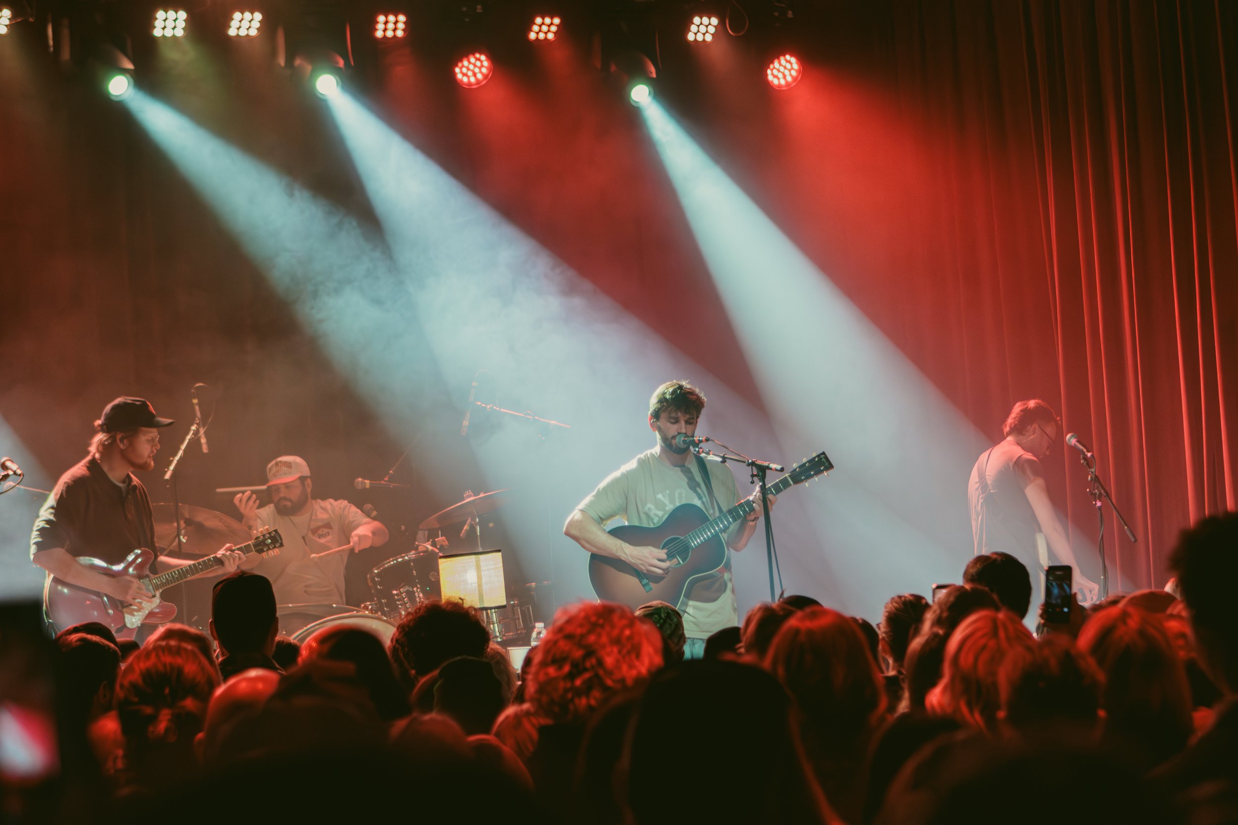 A band performing live on stage with vibrant red and white lighting, including a singer with an acoustic guitar, a guitarist, a drummer, and a keyboardist, in front of an audience.