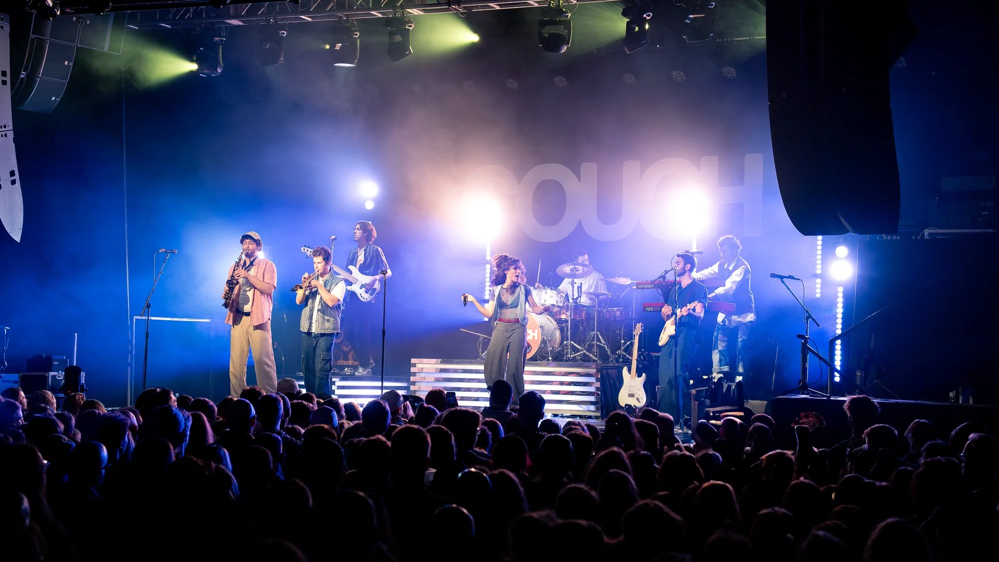 A band performing live on stage with seven members, playing various instruments, under bright stage lights, with an audience in the foreground.