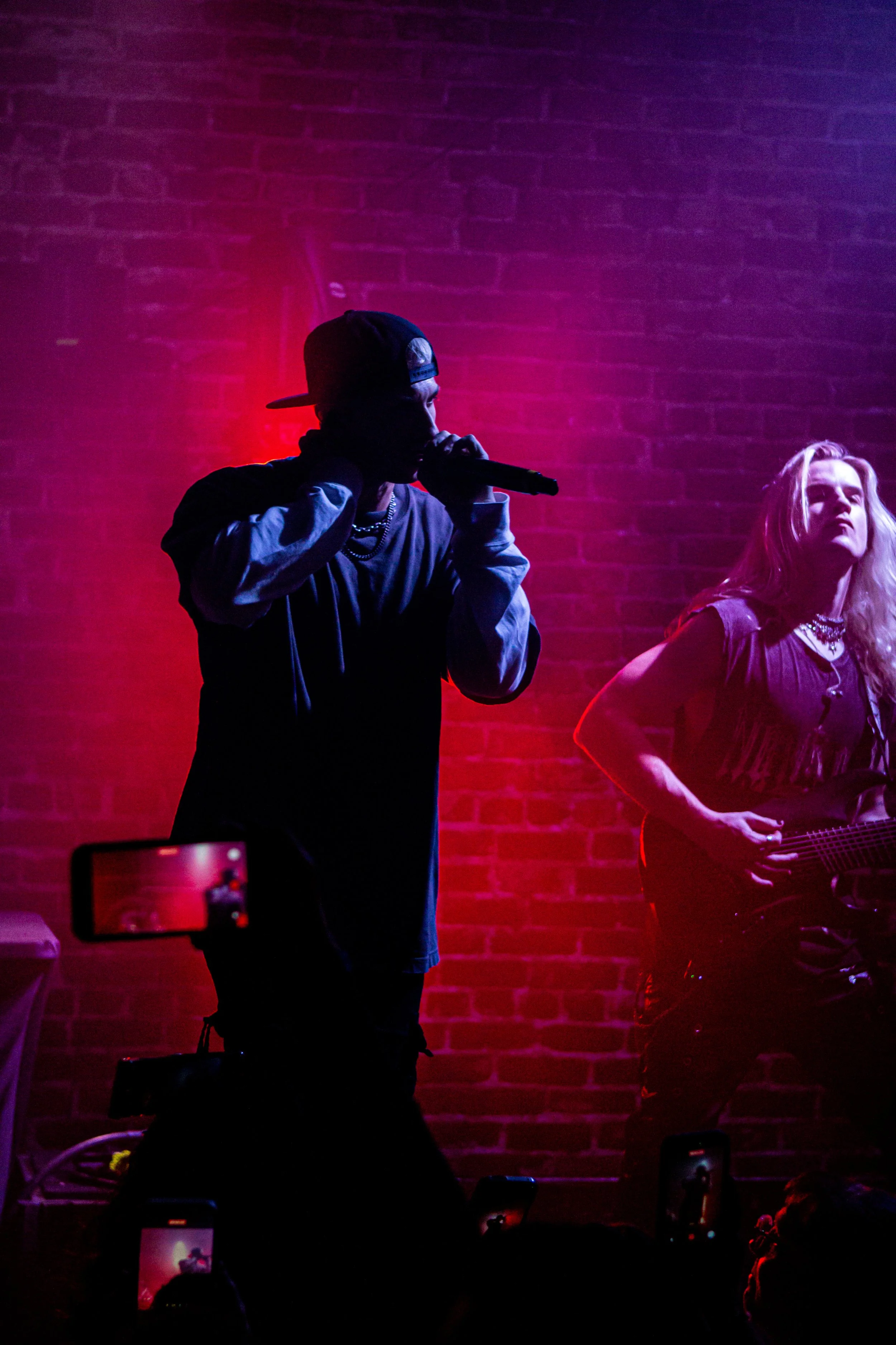A music performance on stage with a male singer in a backward cap and a female guitarist against a brick wall illuminated with pink and purple lighting, with audiences capturing the moment on their phones.