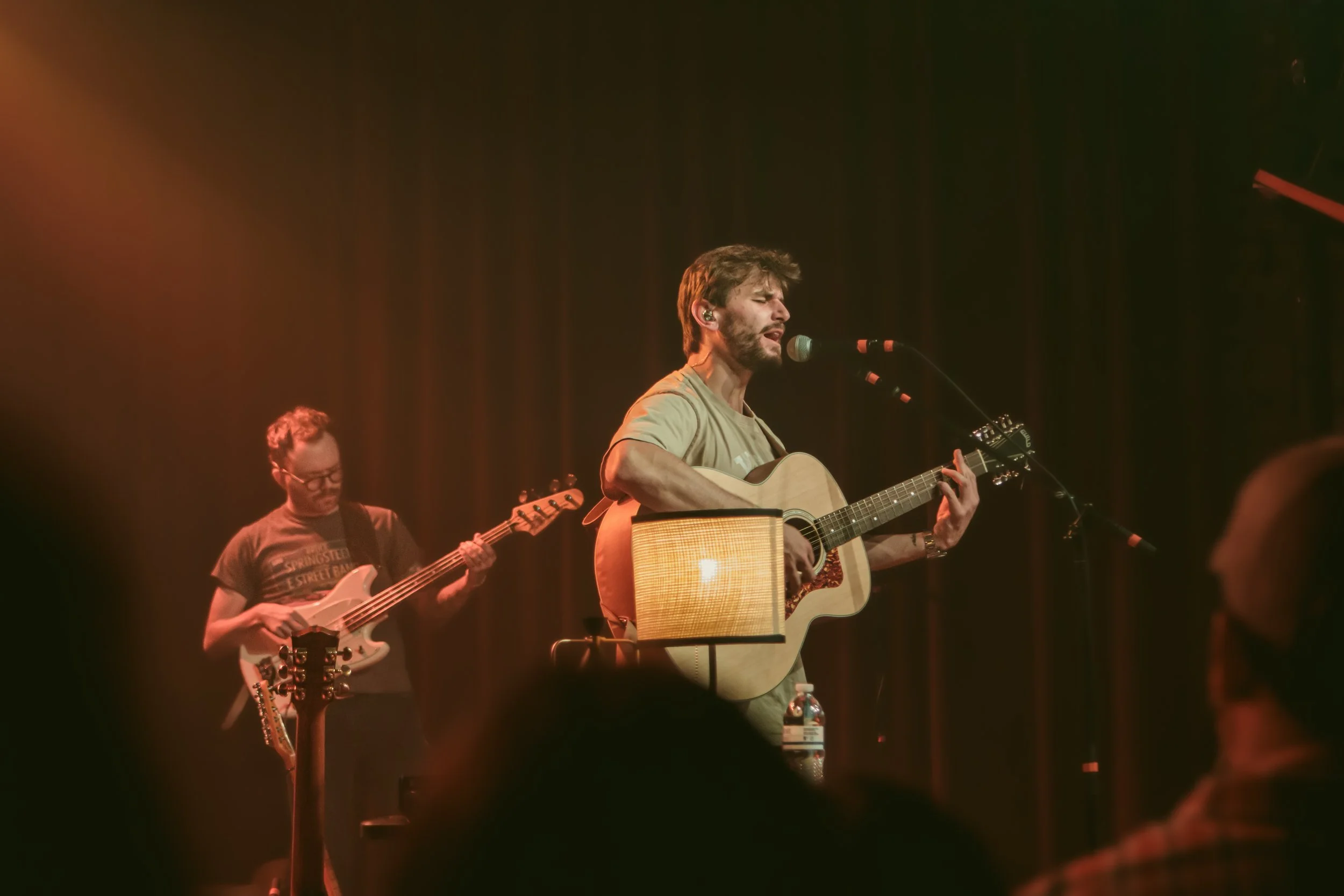 Musicians performing on stage with one playing guitar and singing into a microphone, another playing bass guitar in the background, dim concert lighting, audience silhouettes in the foreground.