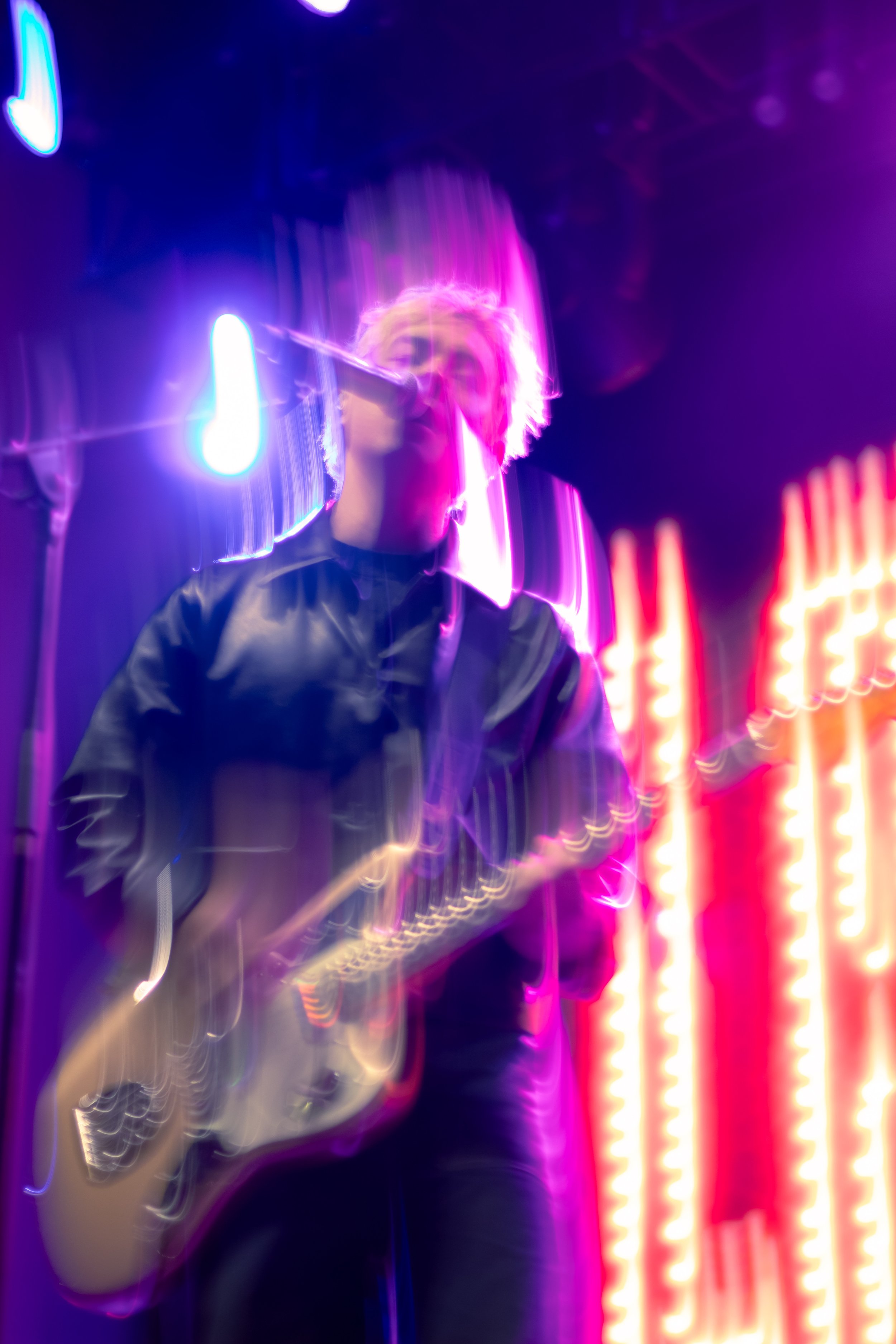 A person with short blonde hair playing an electric guitar on stage with purple and red neon lights.