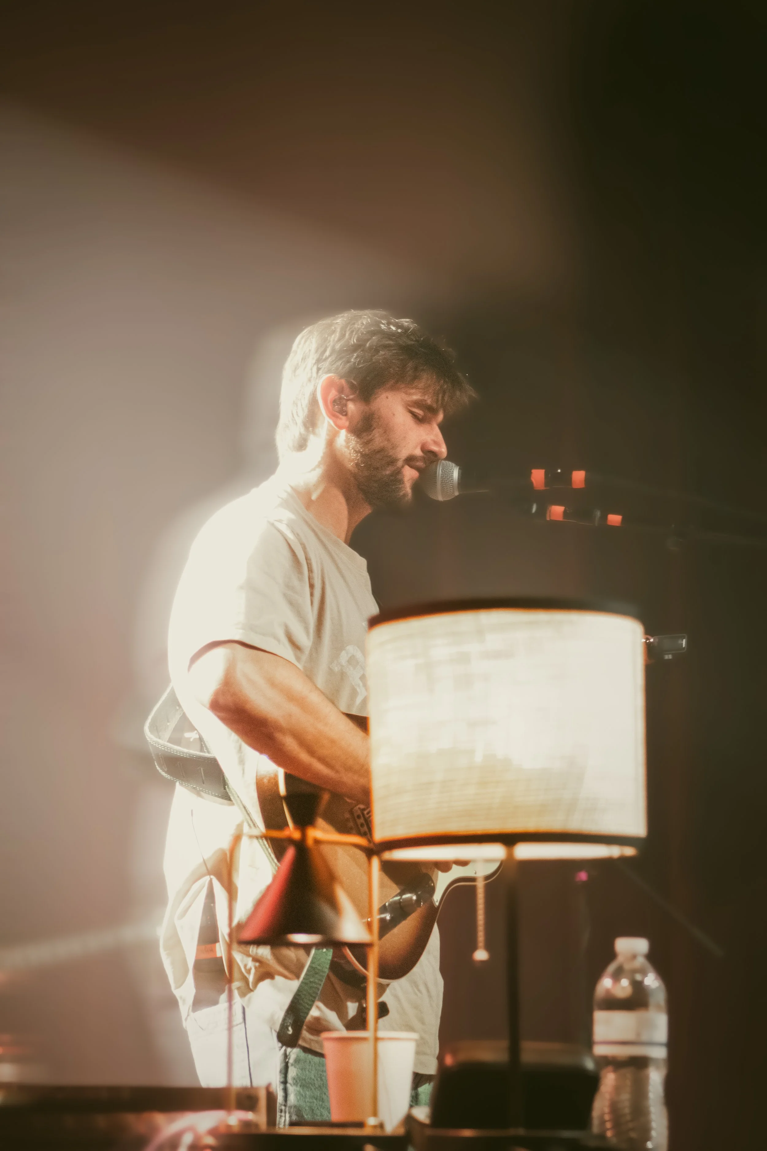 A man with a beard and brown hair performing on stage, playing an instrument and singing into a microphone, illuminated by warm stage lighting.