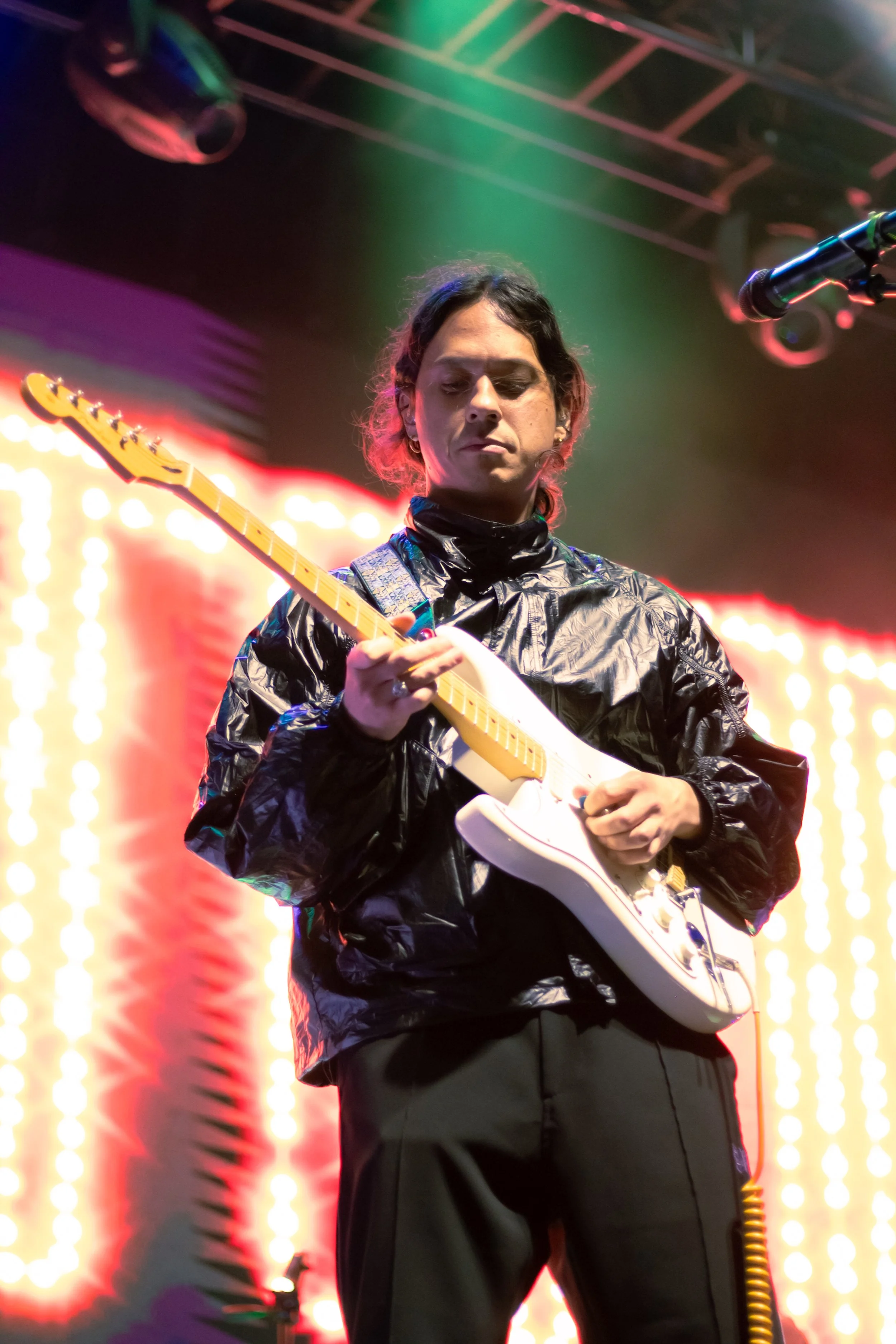 A musician performs on stage, playing a white electric guitar with a yellow neck, wearing a black shiny jacket and black pants, with colorful stage lighting in the background.