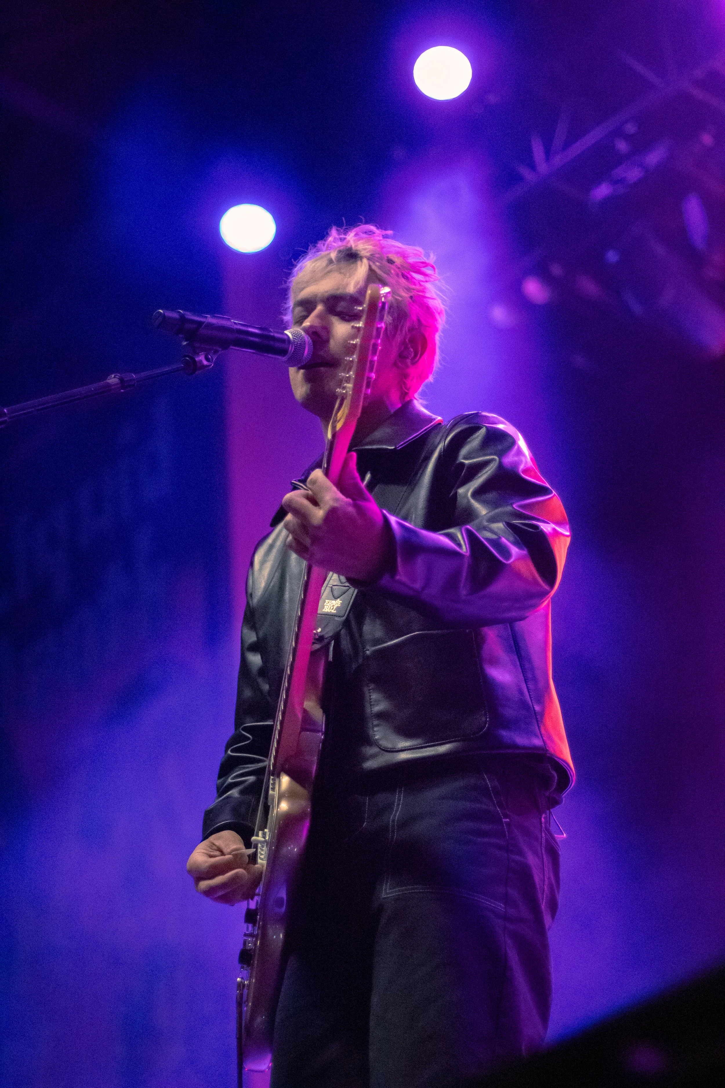 A musician performing on stage, singing into a microphone and playing an electric guitar, under purple and pink stage lighting.