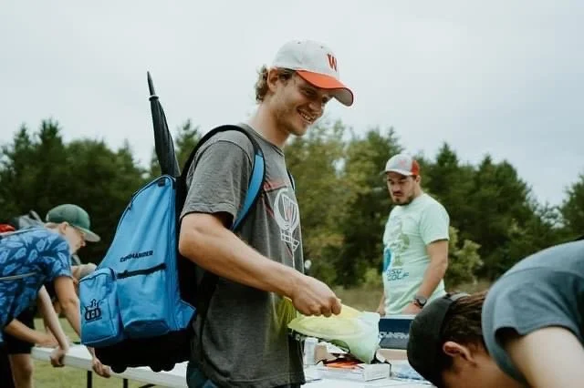 Group of people outdoors at a table, one young man with a backpack smiling, others in the background, trees in the distance.