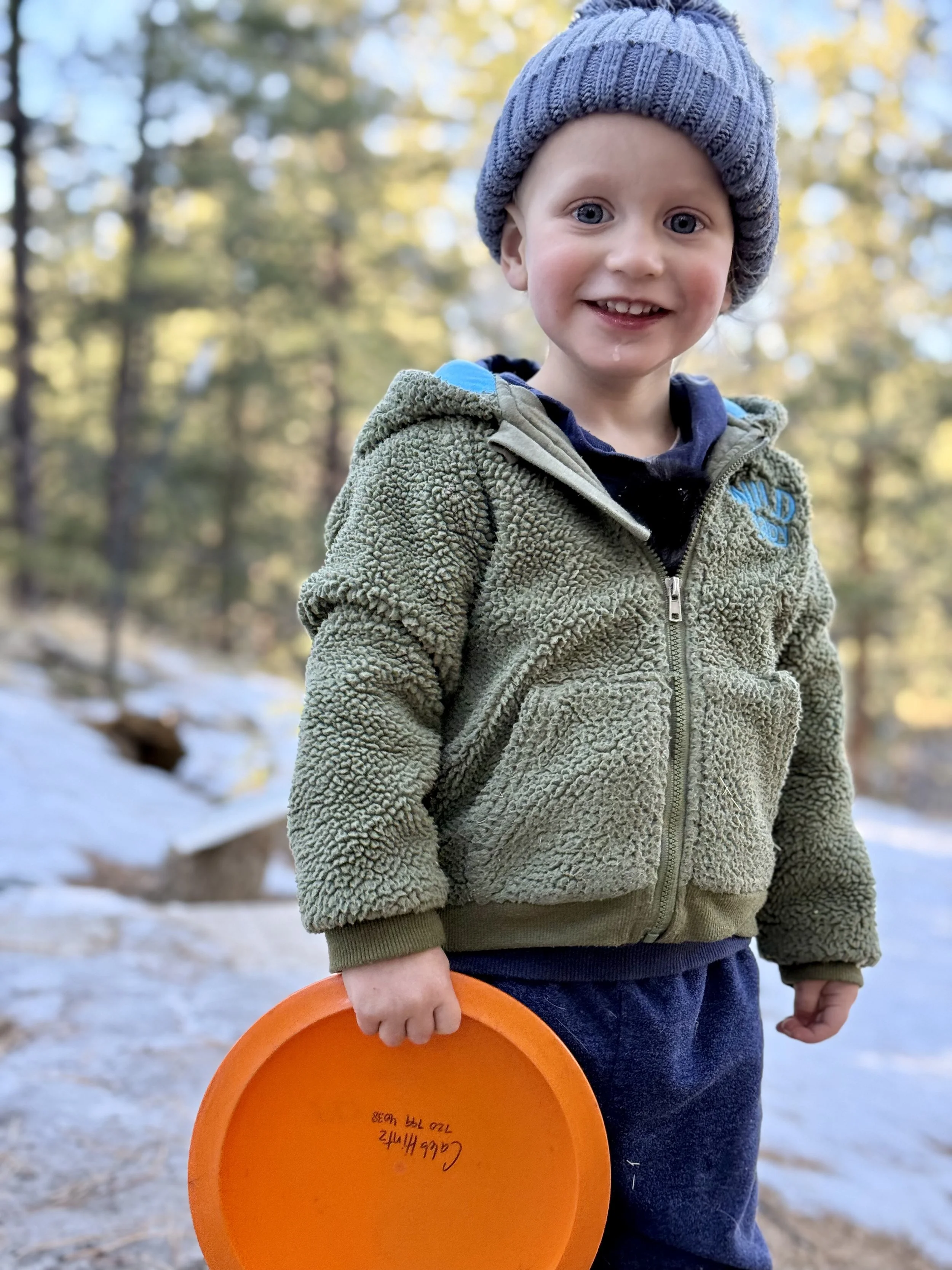 A young boy wearing a gray knit hat and a green fuzzy jacket, standing outdoors on snowy ground in a forest, holding an orange frisbee, smiling at the camera.