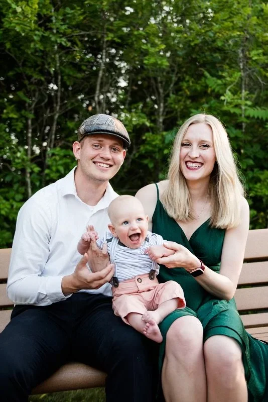 A smiling family sitting on a park bench with a man, woman, and a baby, surrounded by green trees.