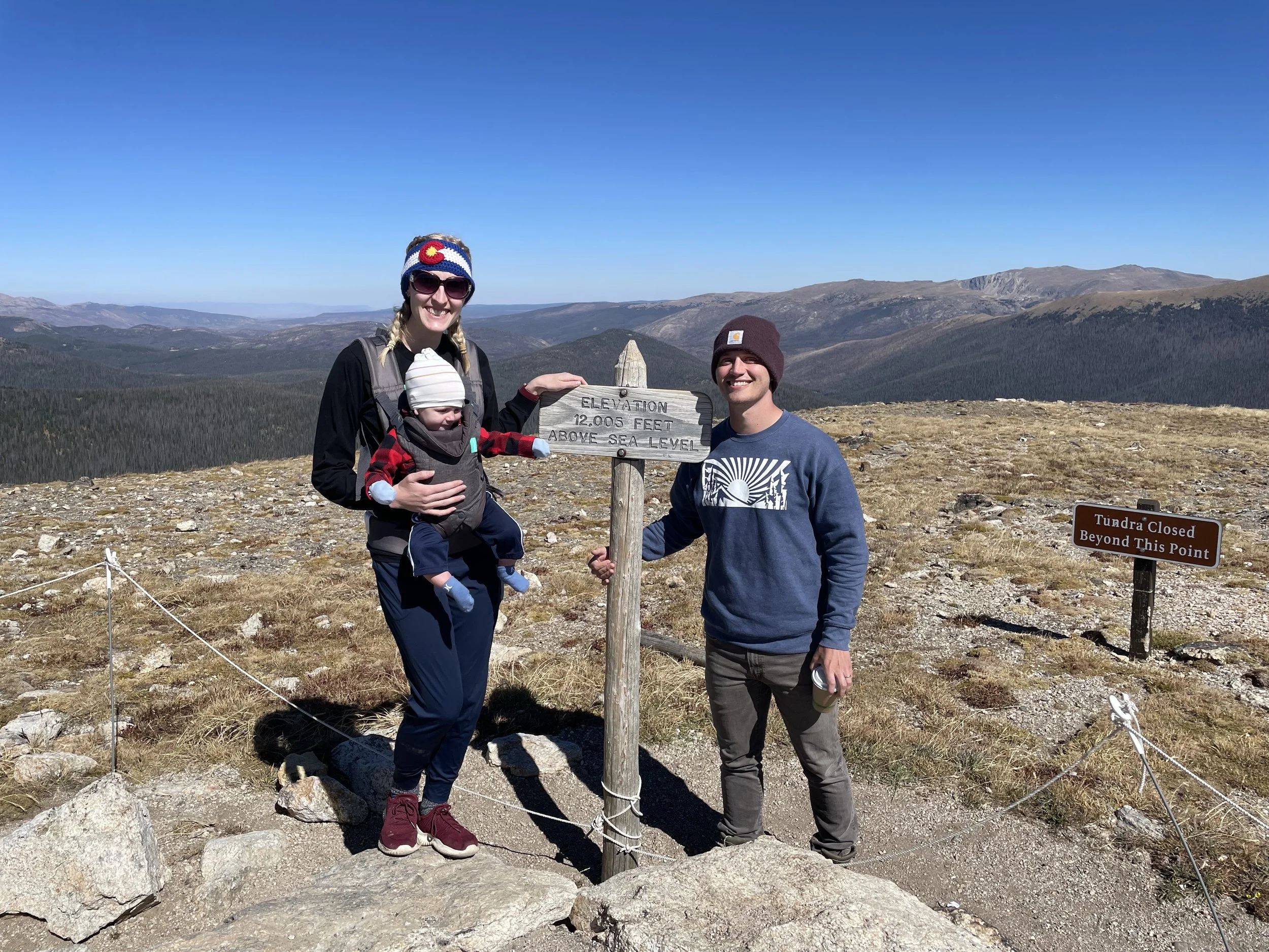 A family of three at the summit of a mountain with a sign indicating an elevation of 12,005 feet above sea level. The woman is holding a child, and the man is standing beside them, smiling. The landscape features mountains and a clear blue sky, and a sign on the right indicates the area is beyond the tundra.