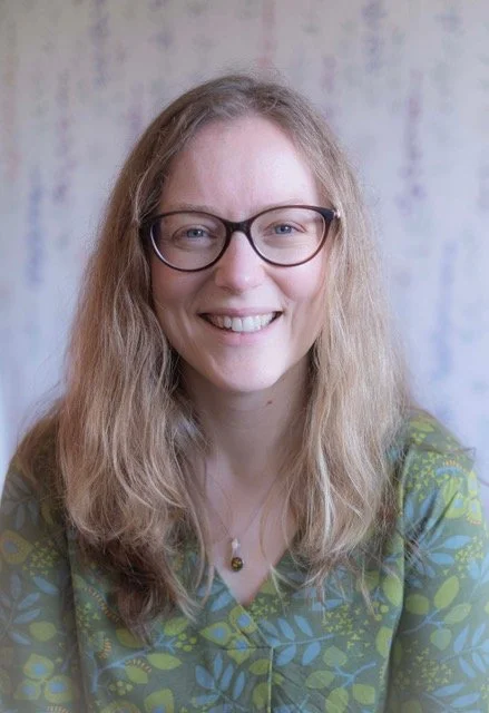 Dr Imogen Marsh, Clinical Psychologist: a woman with long, wavy, light brown hair and glasses, smiling in front of a colorful, blurry background.