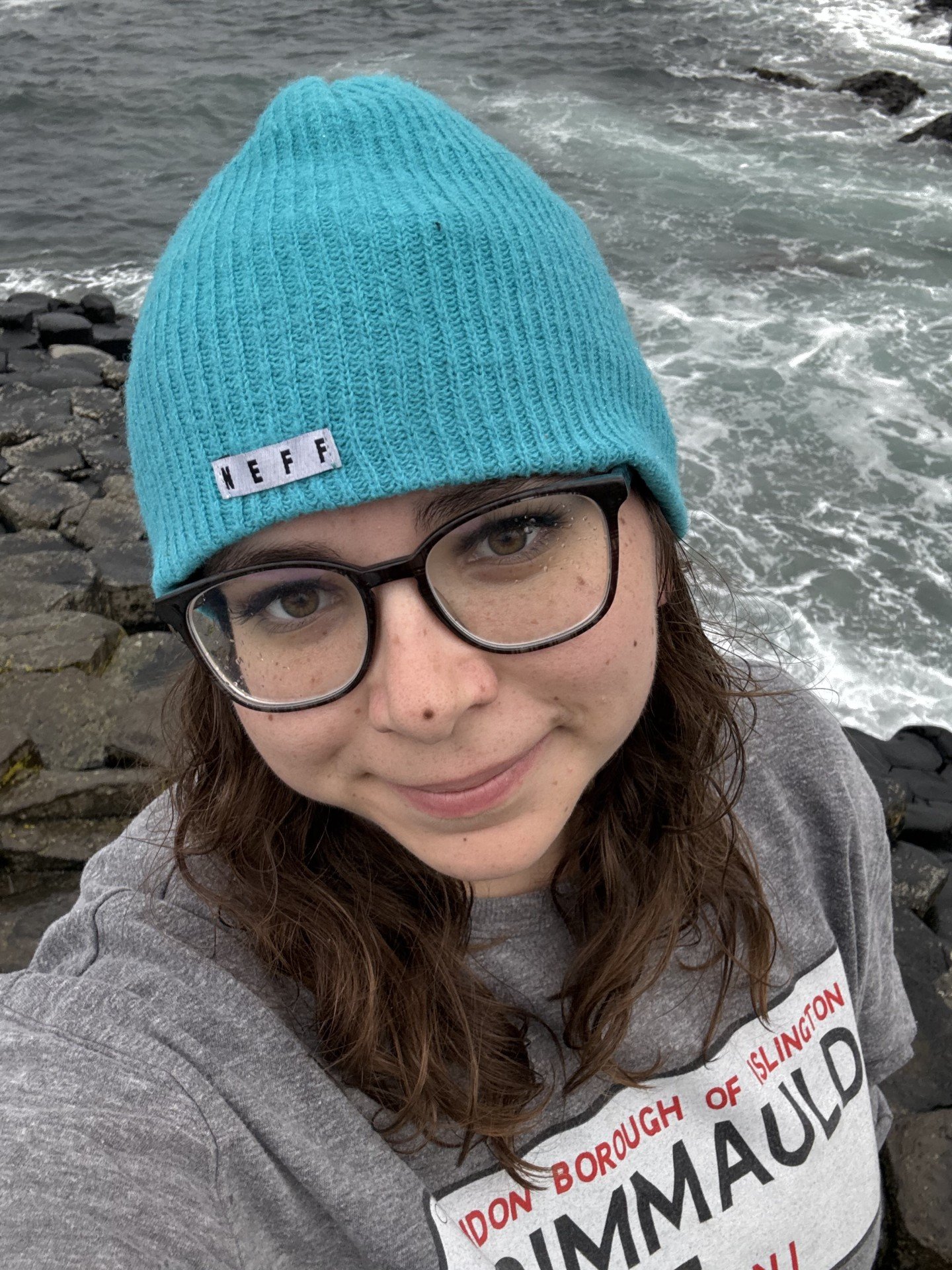 Close-up selfie of a woman wearing glasses and a bright blue knit hat with a 'NEFF' label, standing near rocks and ocean waves.