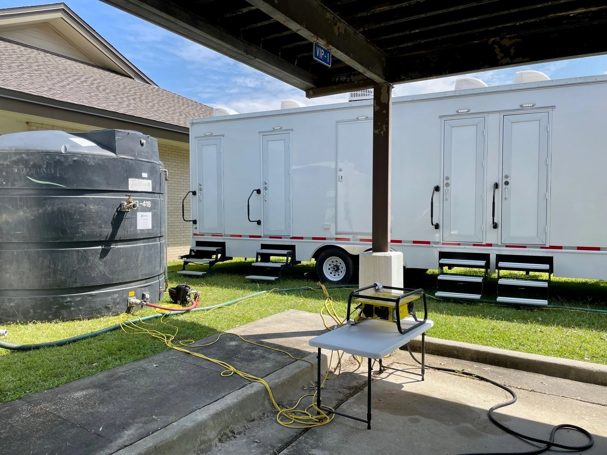 A mobile trailer with a white exterior, black handles on doors, and steps, parked next to a large black water tank, on a grassy area with electrical cords and equipment nearby.