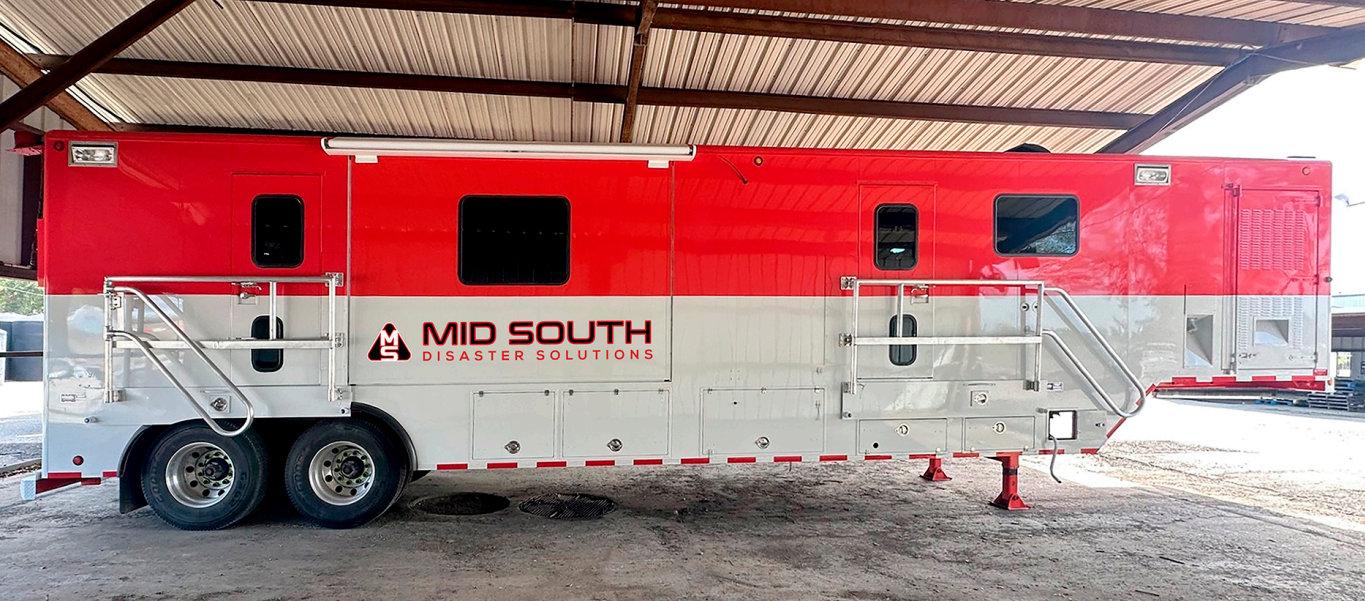 A mobile command center trailer with a two-tone white and blue exterior, parked on a grassy field under a clear blue sky. It has windows, a side door, and a set of stairs leading up to the entrance.