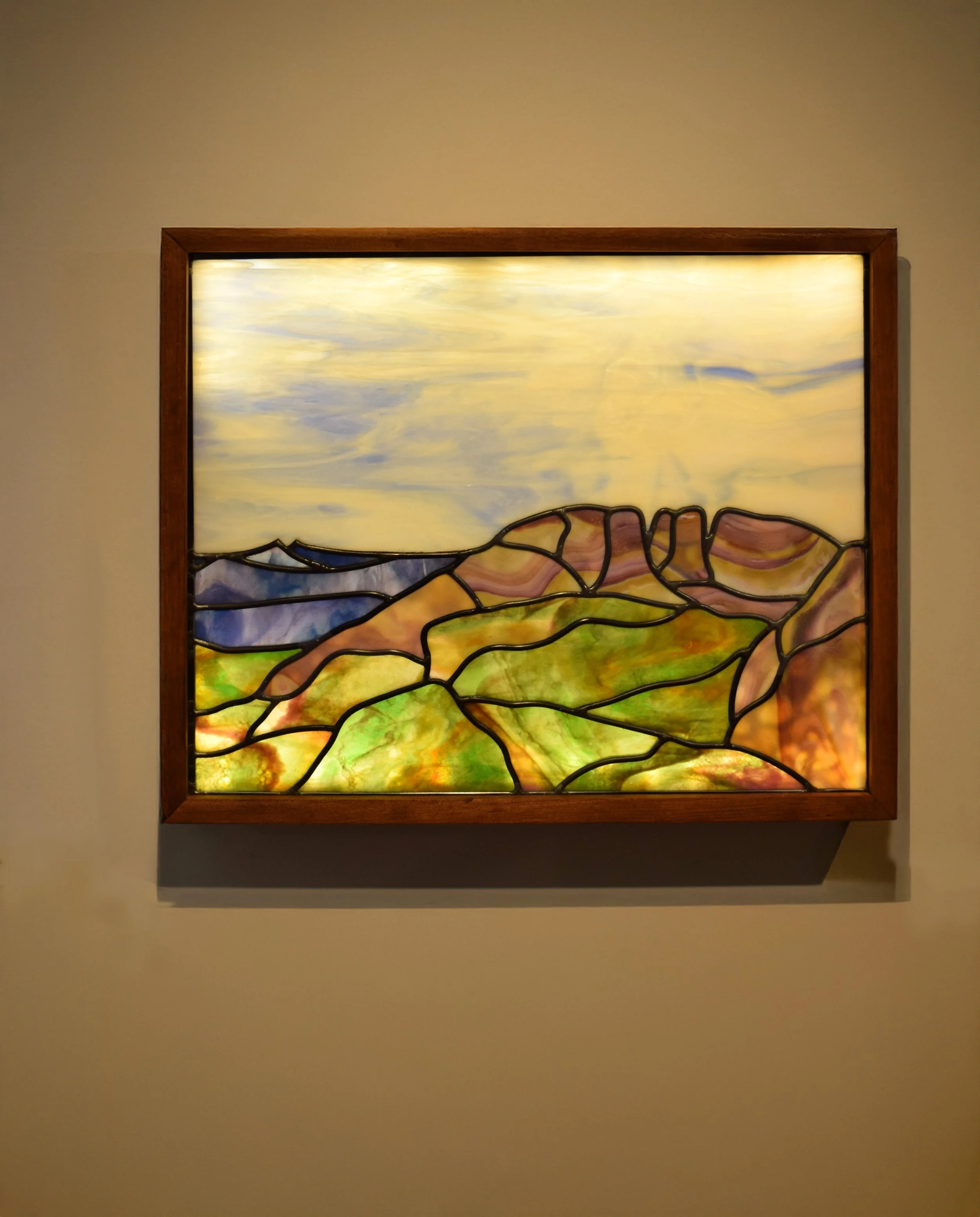 Colorful custom stained glass window with a mountain landscape scene, framed in wood, mounted on a beige wall with soft lighting.