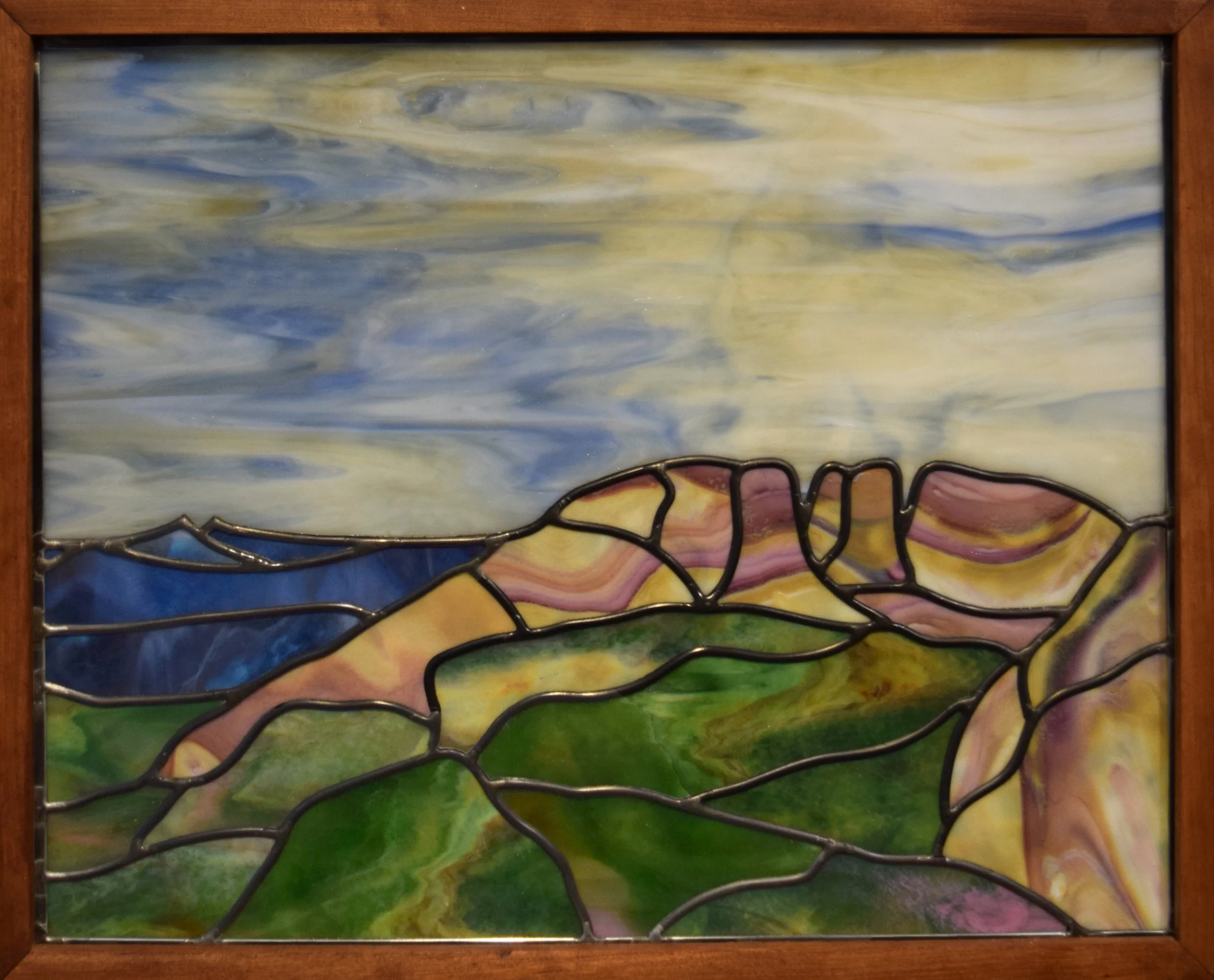 Colorful custom stained glass window with an abstract landscape scene featuring green hills, a blue sky, and wispy clouds, framed in wood.