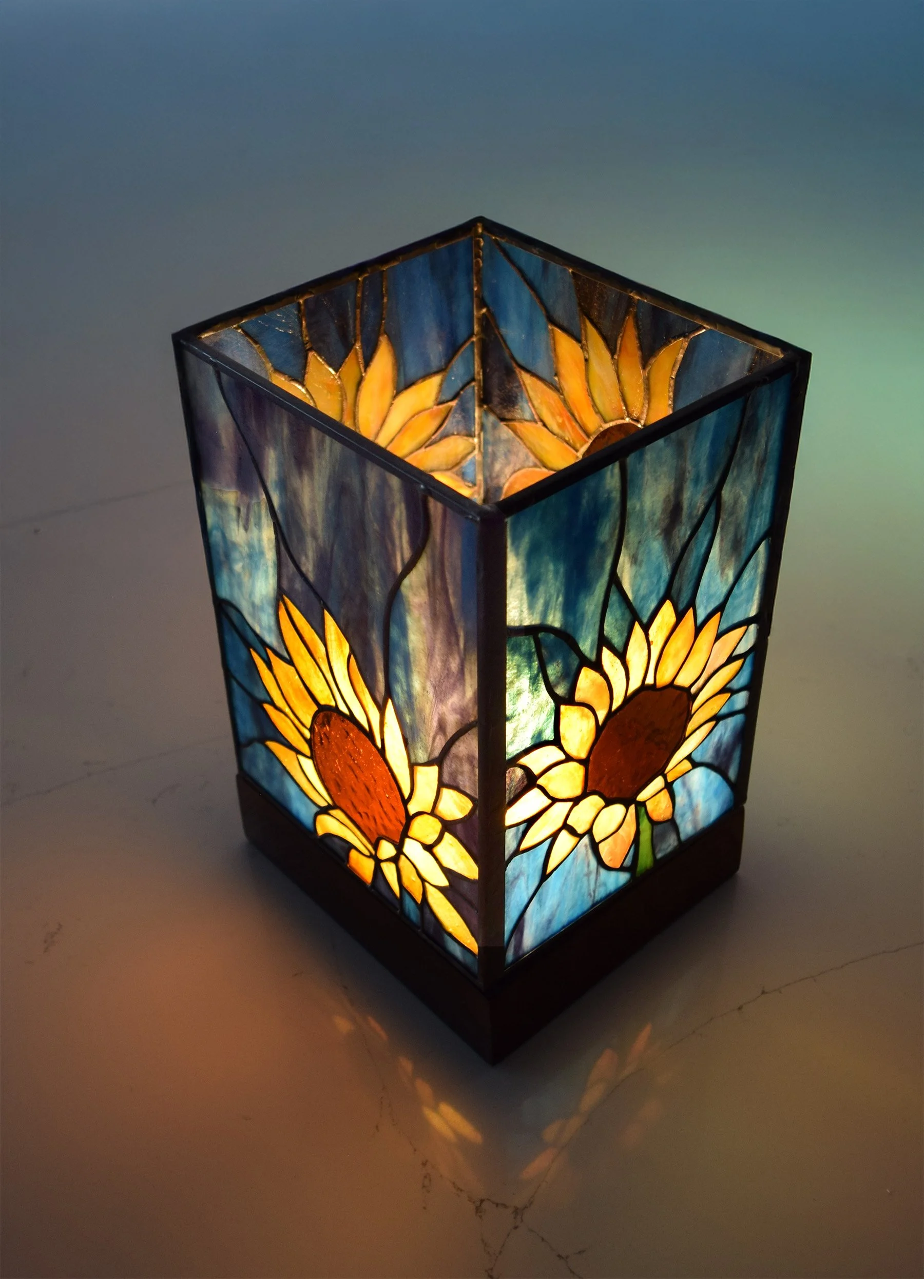 A stained glass lamp featuring sunflower designs with yellow petals and brown centers, illuminated from within, casting a glow on the surface below.