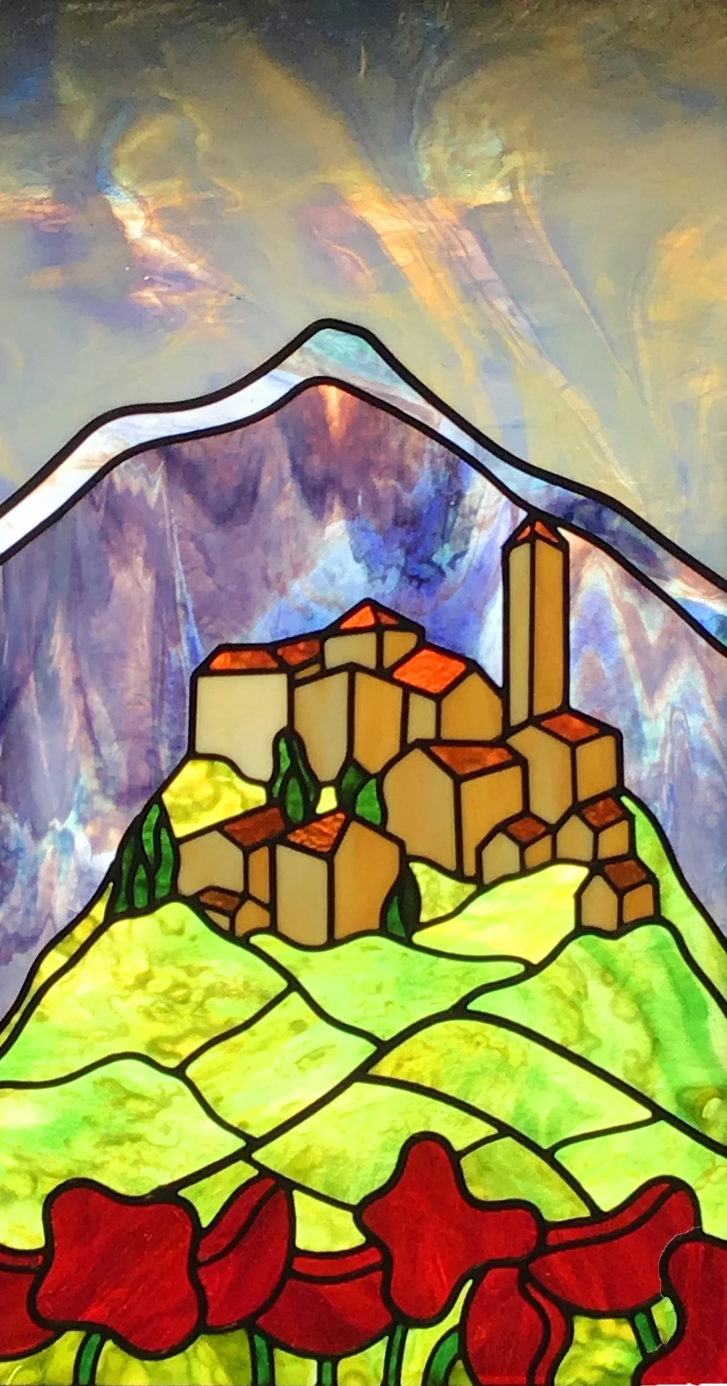 Colorful stained glass window depicting a mountain landscape with a village on a hillside, surrounded by greenery and red flowers at the bottom.