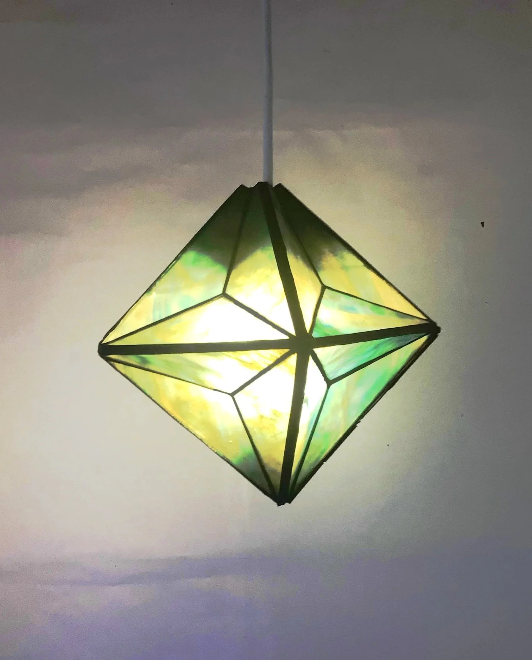 A geometric, stained-glass style hanging light fixture with yellow and green hues, illuminated against a plain background.