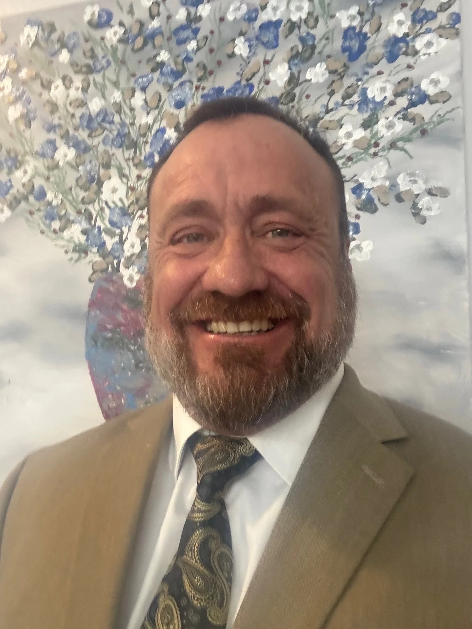 A man in a tan suit and patterned tie smiling in front of a painting of a blooming tree with white and blue flowers.
