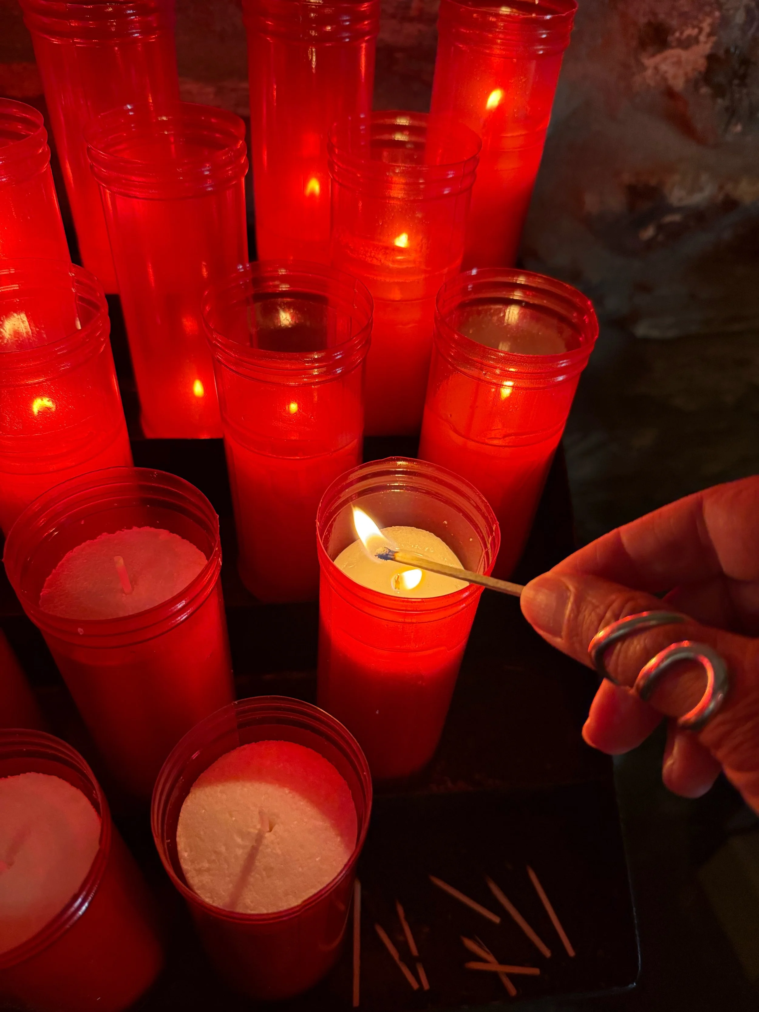 A person's hand lighting a white candle in a red candle holder with a matchstick, surrounded by several other red candles, some lit and some unlit.