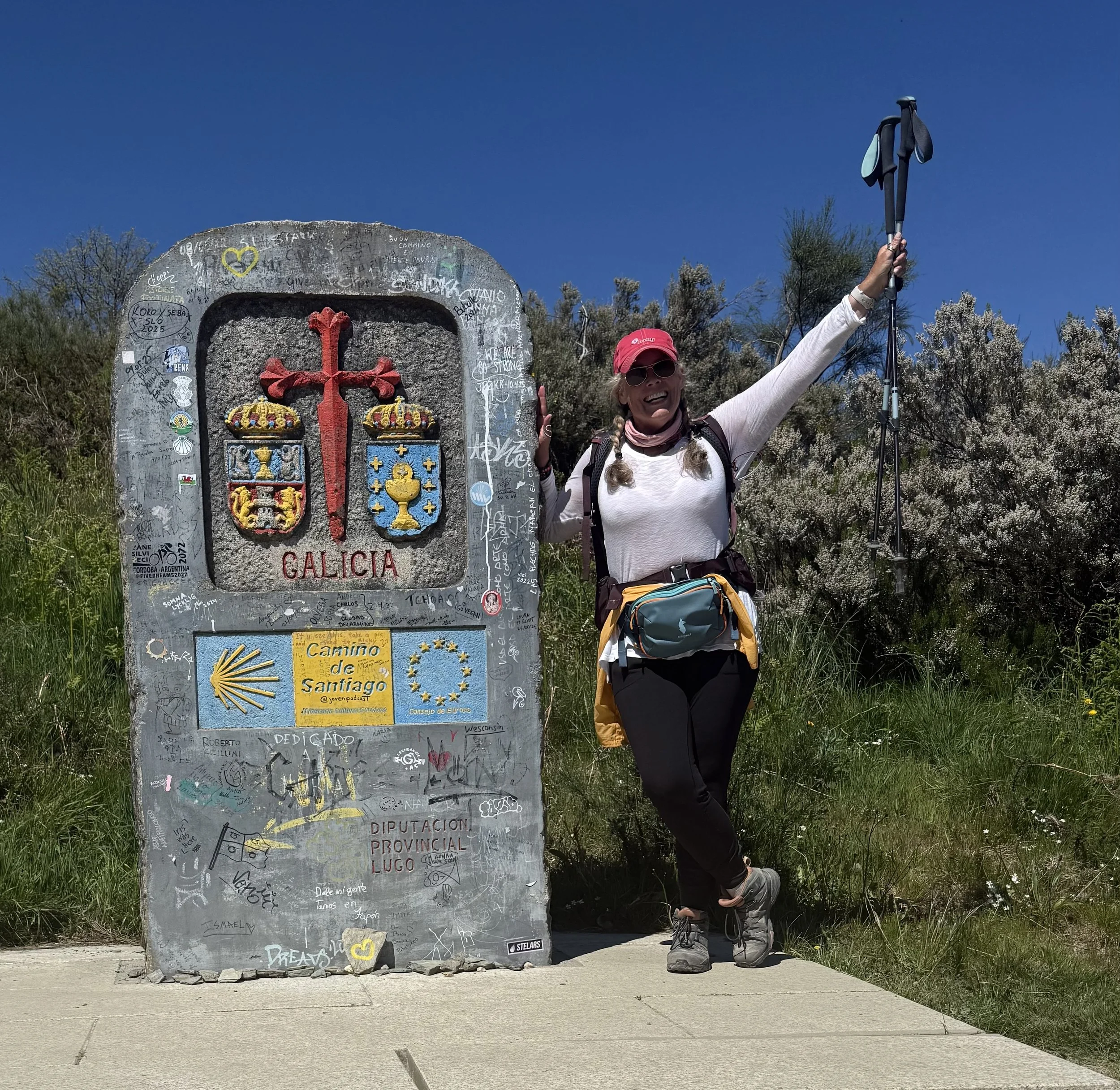 Woman with a pink hat, sunglasses, and a hiking backpack standing next to a stone marker with the Galicia coat of arms, stickers, and graffiti. She is holding trekking poles and smiling, with lush bushes and a clear blue sky in the background.