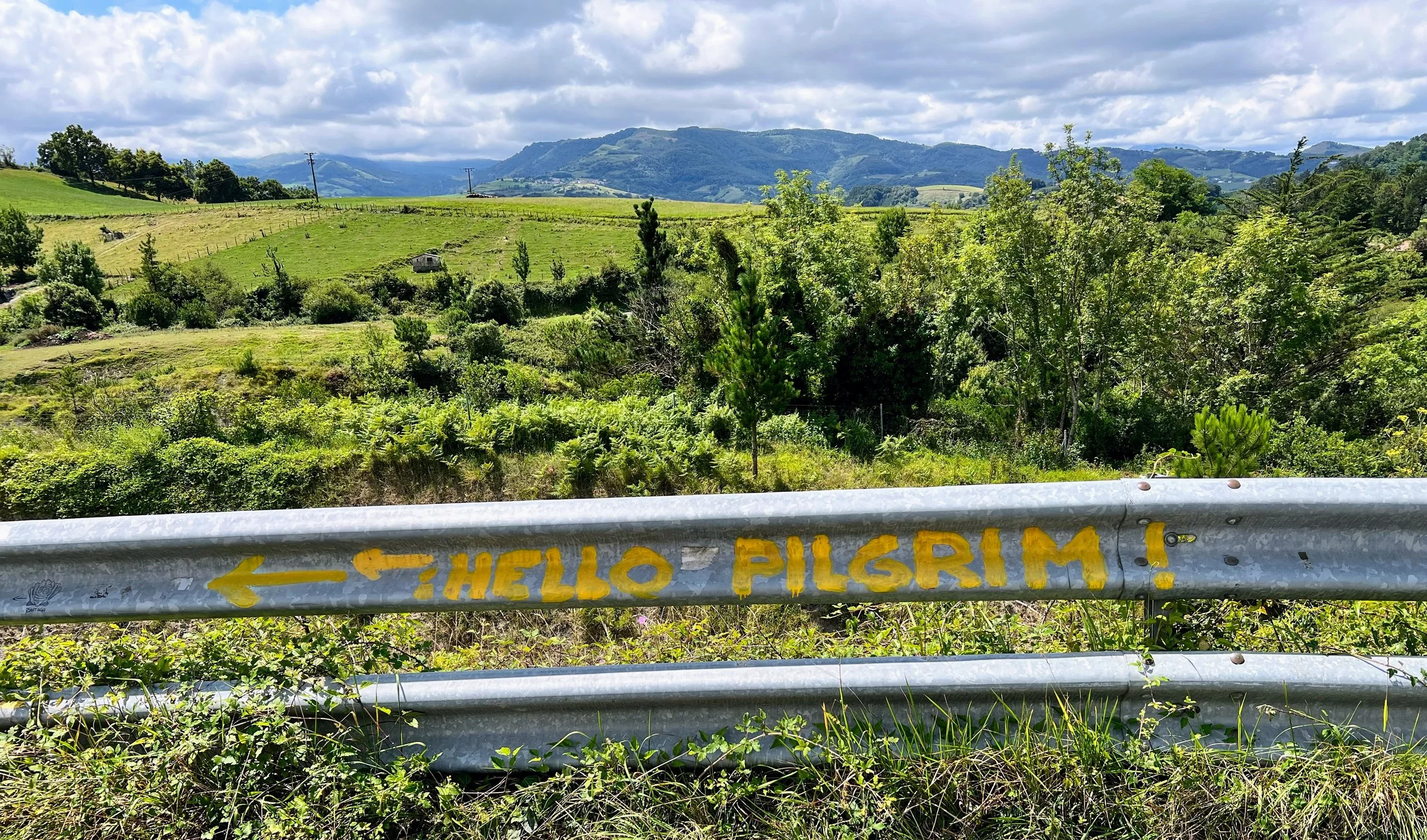 Graffiti on a metal guardrail that says 'HELLO PILGRIM' with an arrow pointing left, set against a lush green countryside with rolling hills, trees, and a partly cloudy sky.