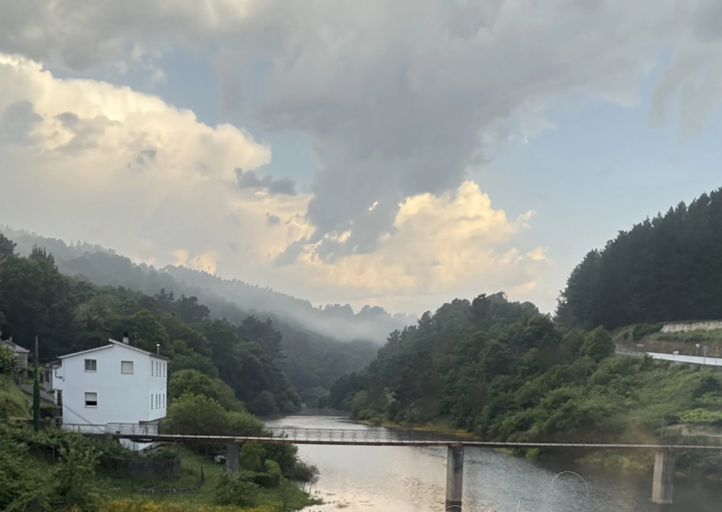 A river flowing through a lush, green valley with hills covered in trees on either side. A white, three-story house and a bridge crossing the river are visible in the foreground. The sky is cloudy with patches of blue, and mist is seen over the distant hills.