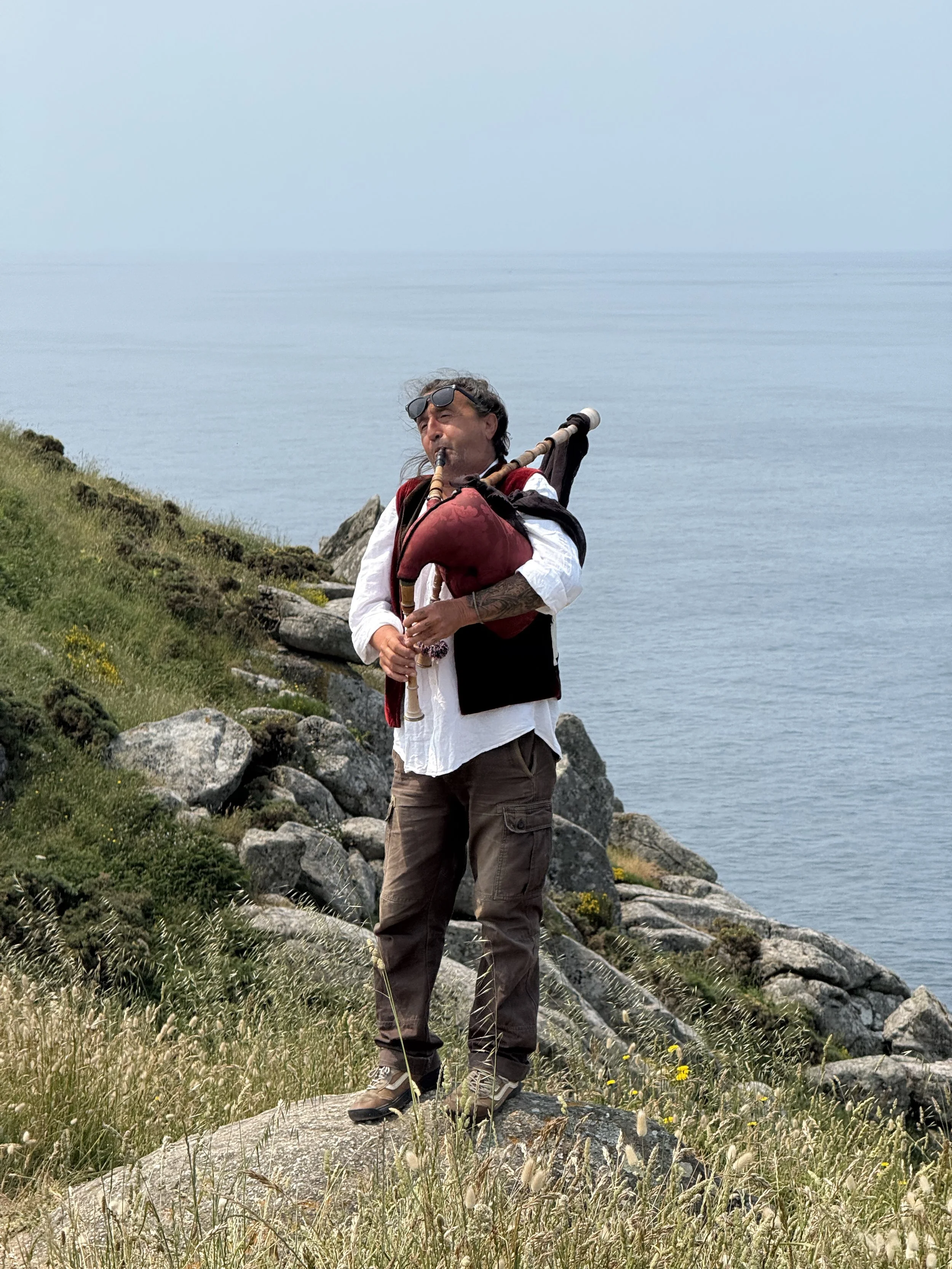 A man standing on a grassy and rocky cliff playing bagpipes near the ocean.