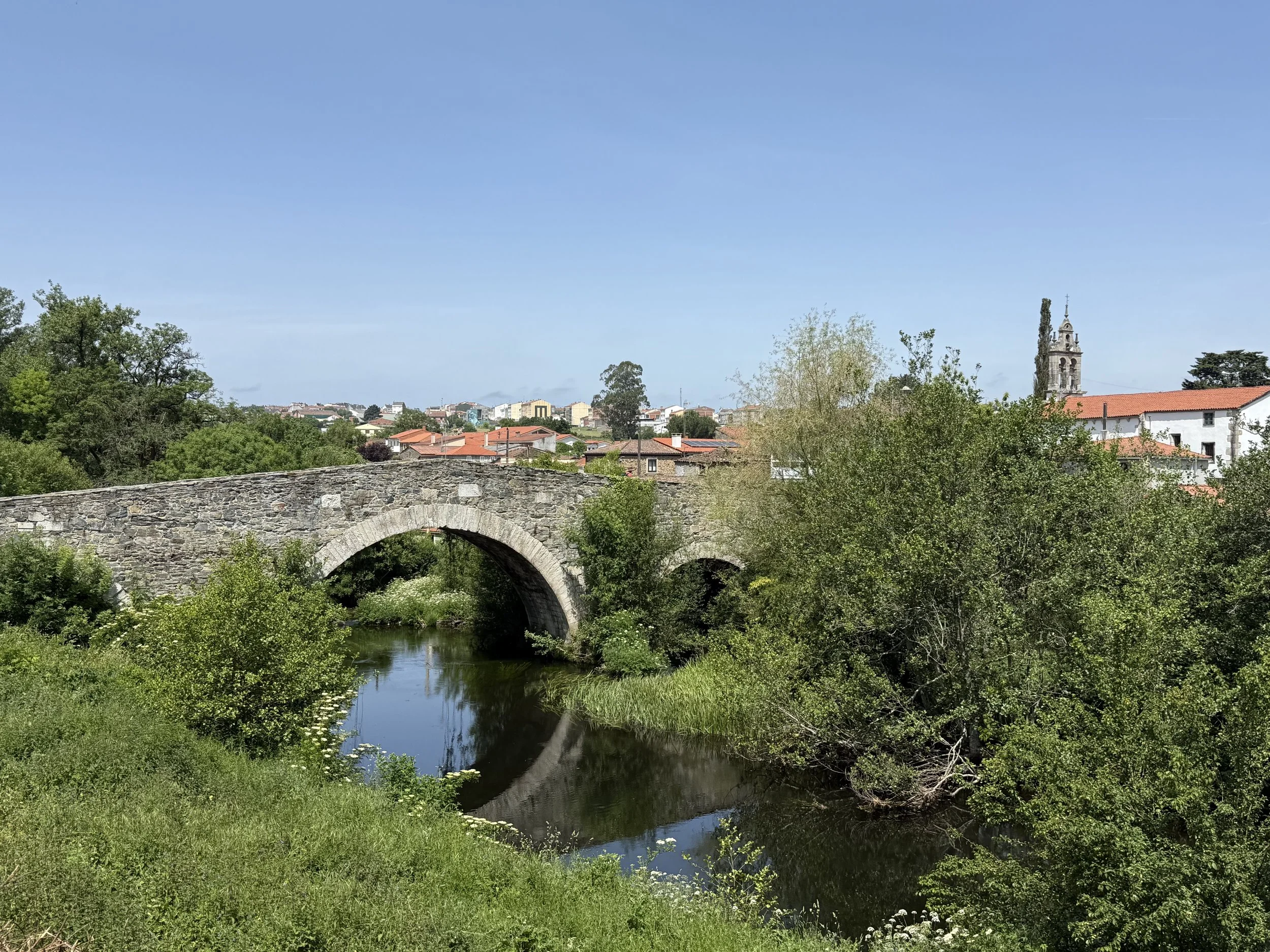A stone bridge crosses a small river surrounded by green trees and grass, with a town featuring red-tiled roofs and a church tower in the background under a clear blue sky.