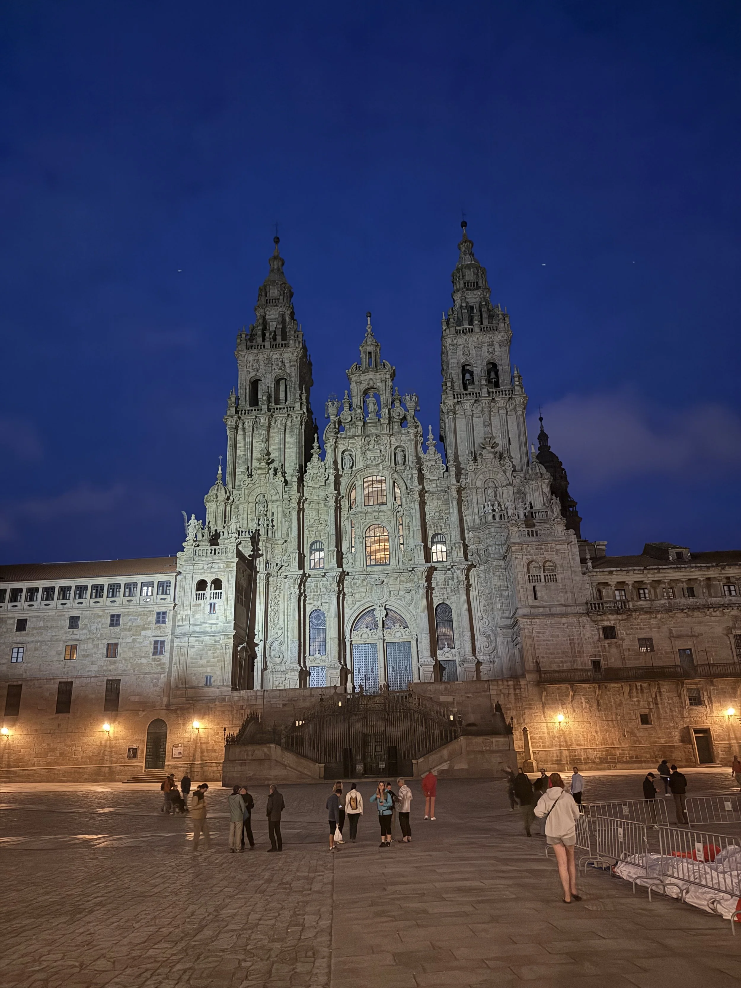 Night view of a large historic cathedral with two tall towers, lit up in the evening, with people gathered in front of it.