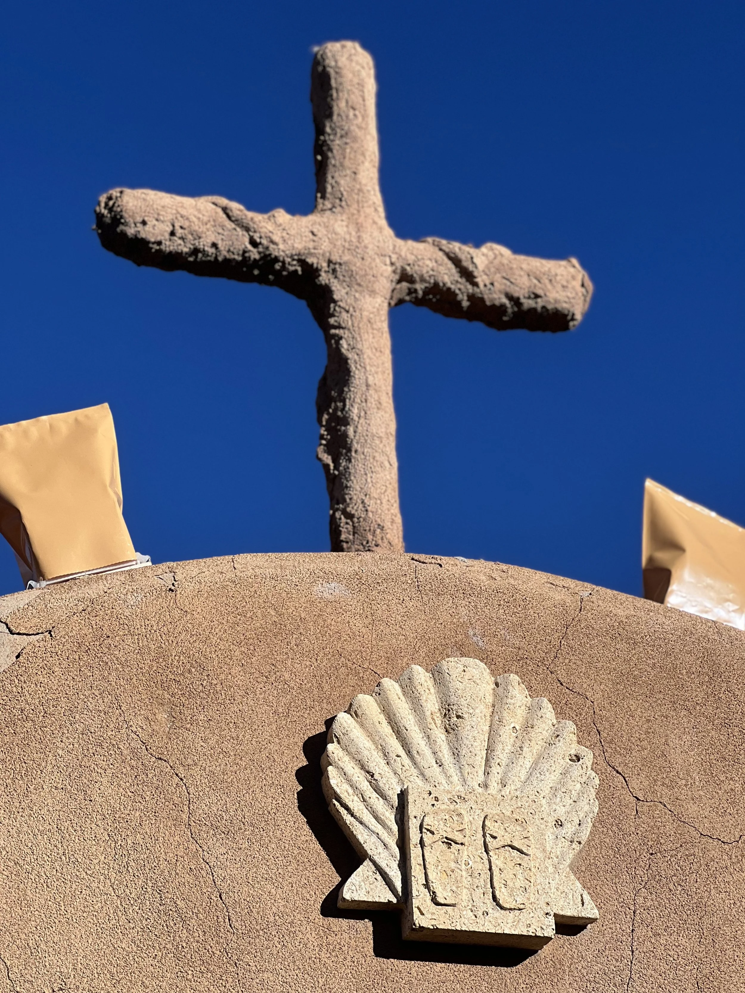 Close-up of a stone cross on top of a building with a board and a stone shell emblem in front of it, against a clear blue sky.