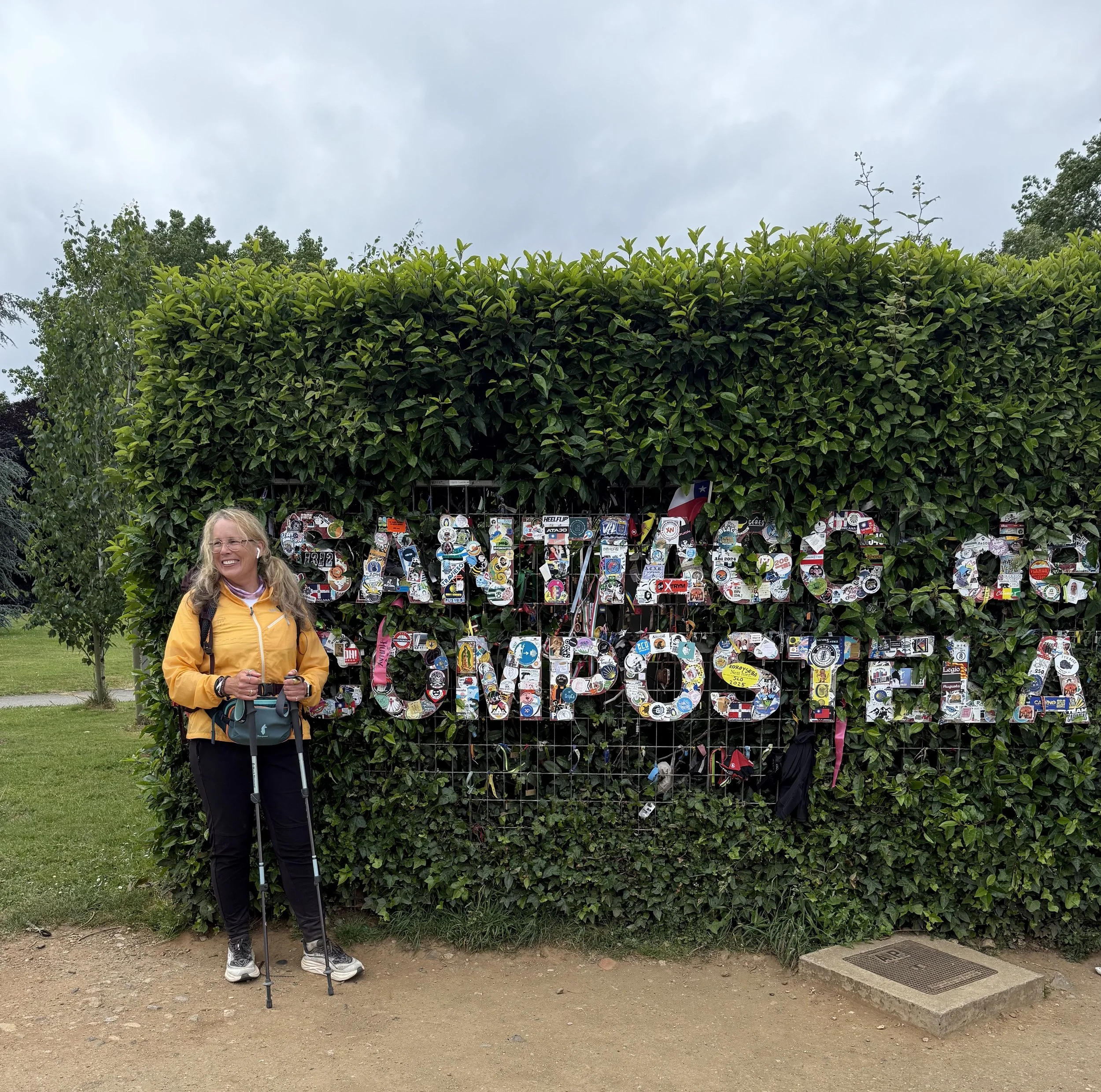 A woman standing next to a metal grid with a large sign spelling 'GALVESTON' decorated with colorful stickers, in front of a bushy background and cloudy sky.