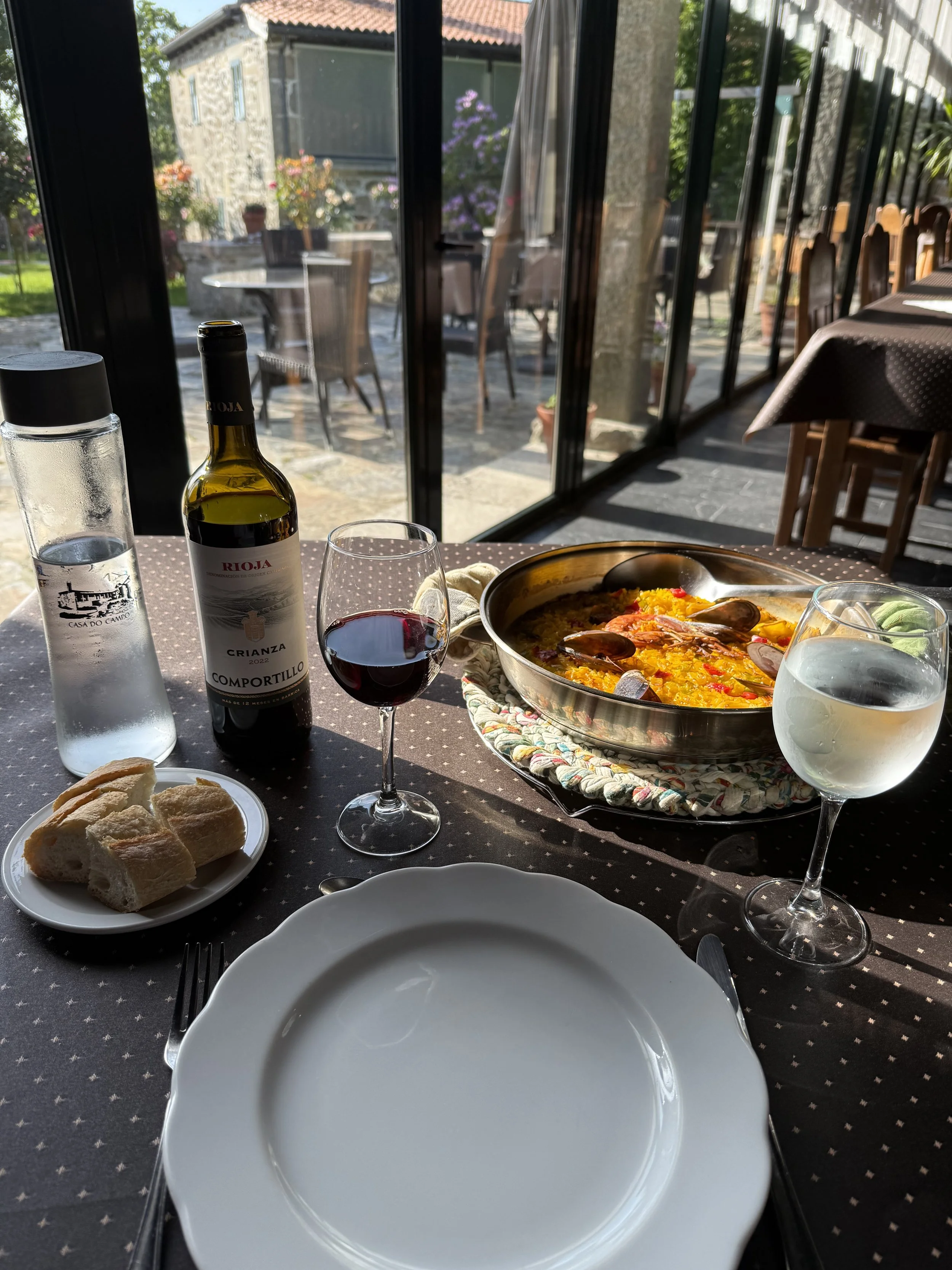 A table set with a large pot of paella, a glass of red wine, a glass of white wine, a bottle of Rioja wine, a plate with bread rolls, a water glass, and tableware, with a view of an outdoor patio area through glass doors.