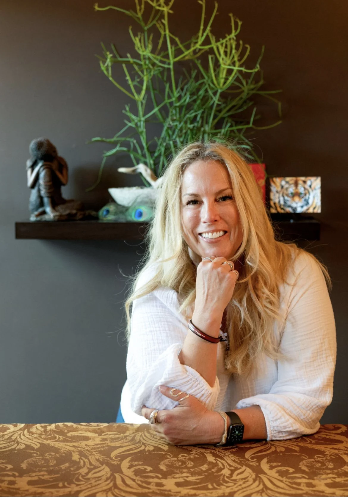 Lucinda Dillon, smiling and resting her chin on her right hand, seated at a table with a patterned tablecloth, with a dark wall, green plant, decorative sculpture, peacock feather ornament, and a tiger face artwork in the background.