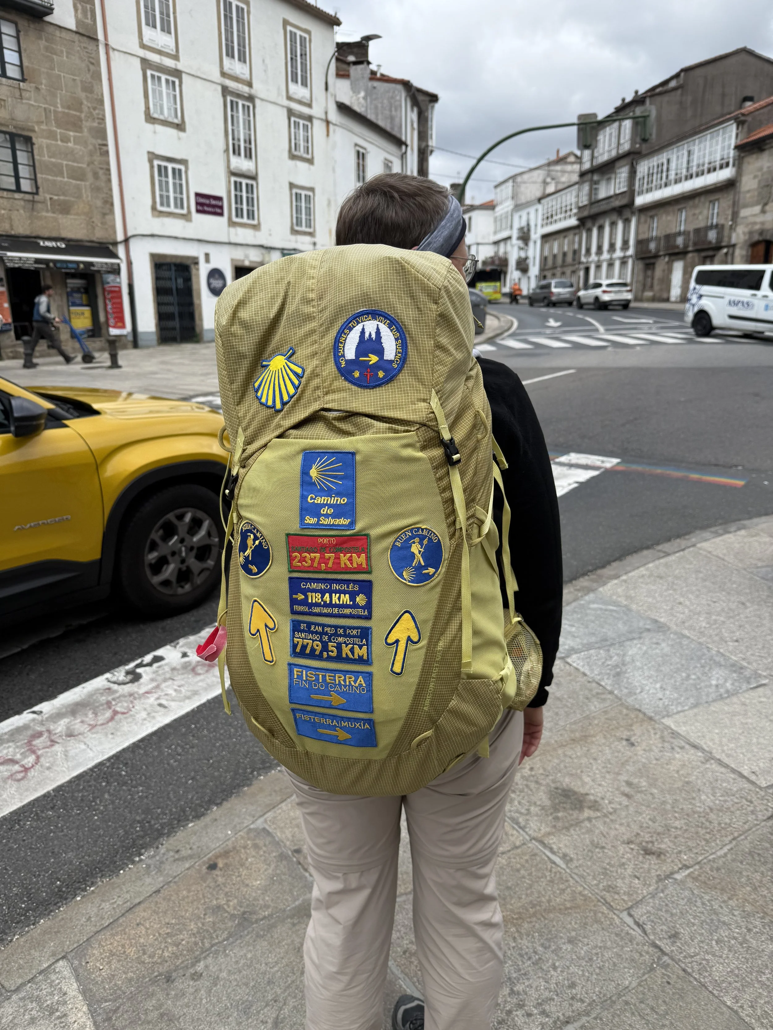 Person with short hair, glasses, and a headband walking on a city sidewalk with a large yellow backpack covered in patches and icons.